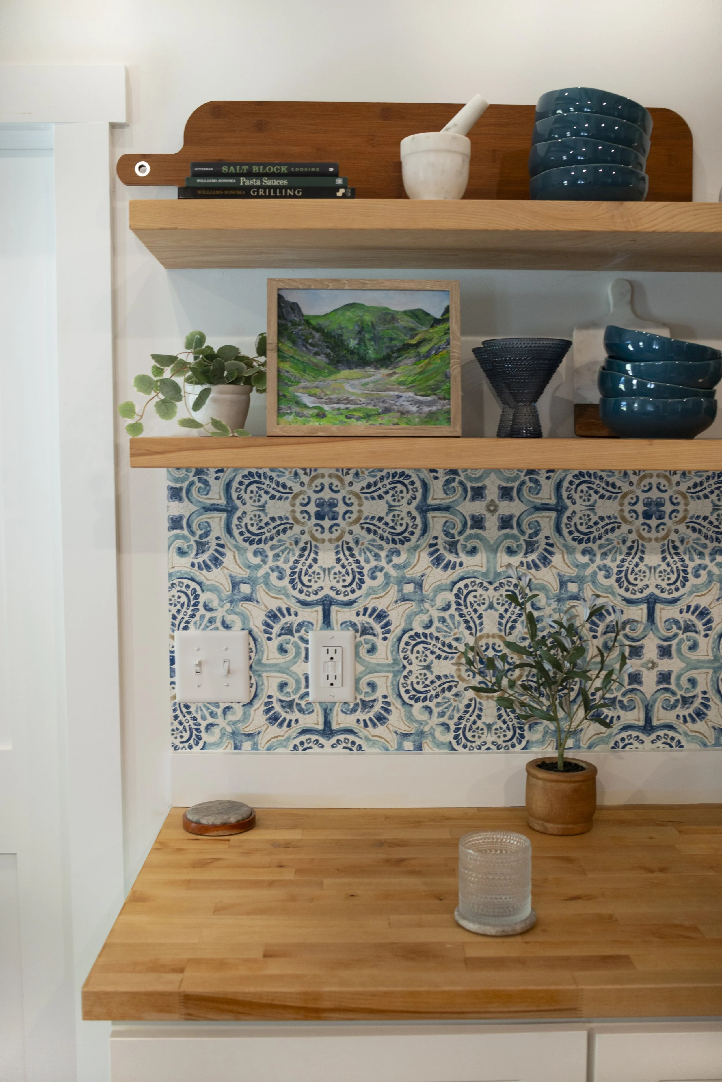 A print of Glencoe in the Scottish Highlands in a wooden frame displayed on a wooden shelf in a cozy, modern kitchen with colorful blue accents.