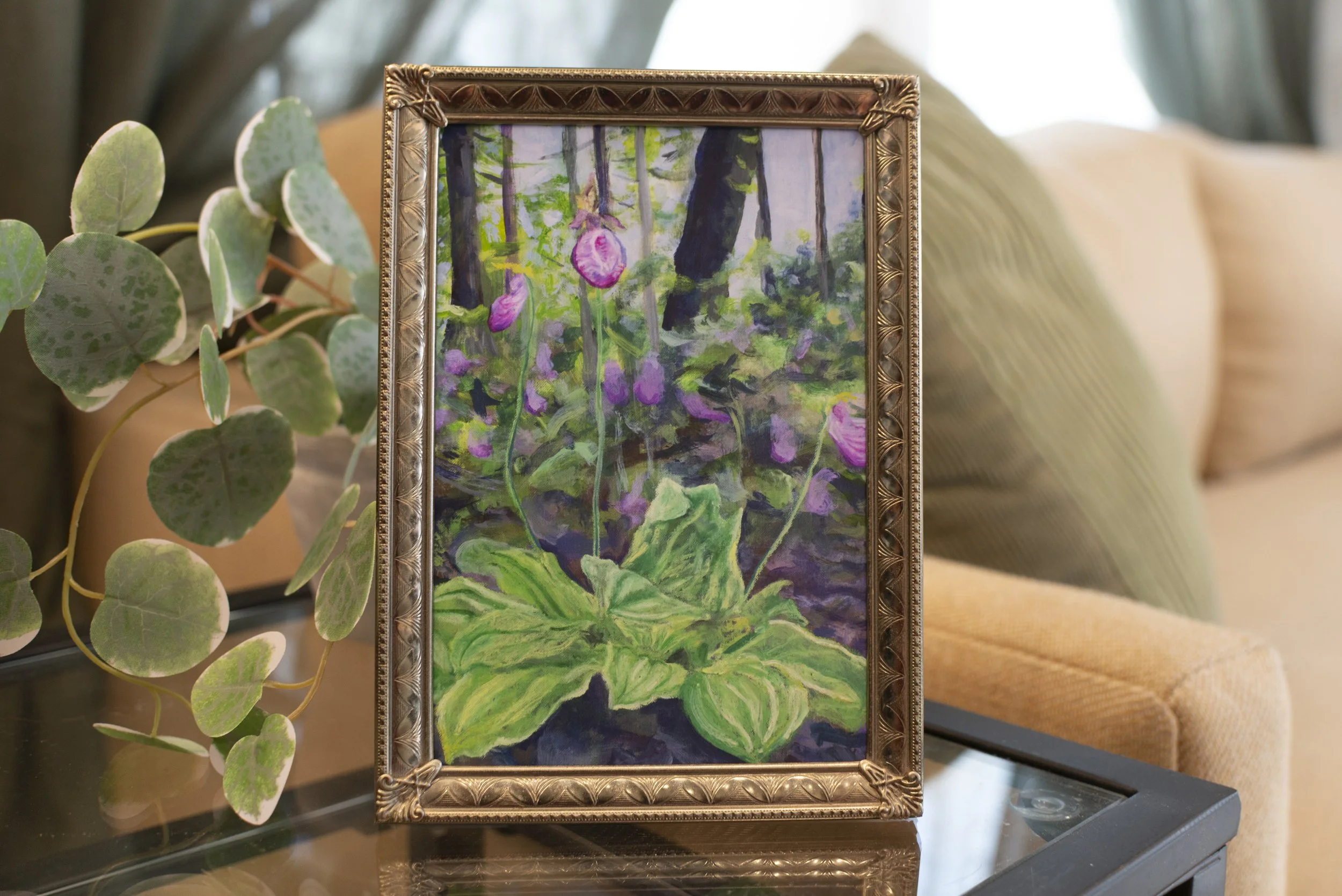 A print of pink Lady Slipper Orchids in a gold frame, displayed on a side table in a living room next to a cozy couch and indoor plant.