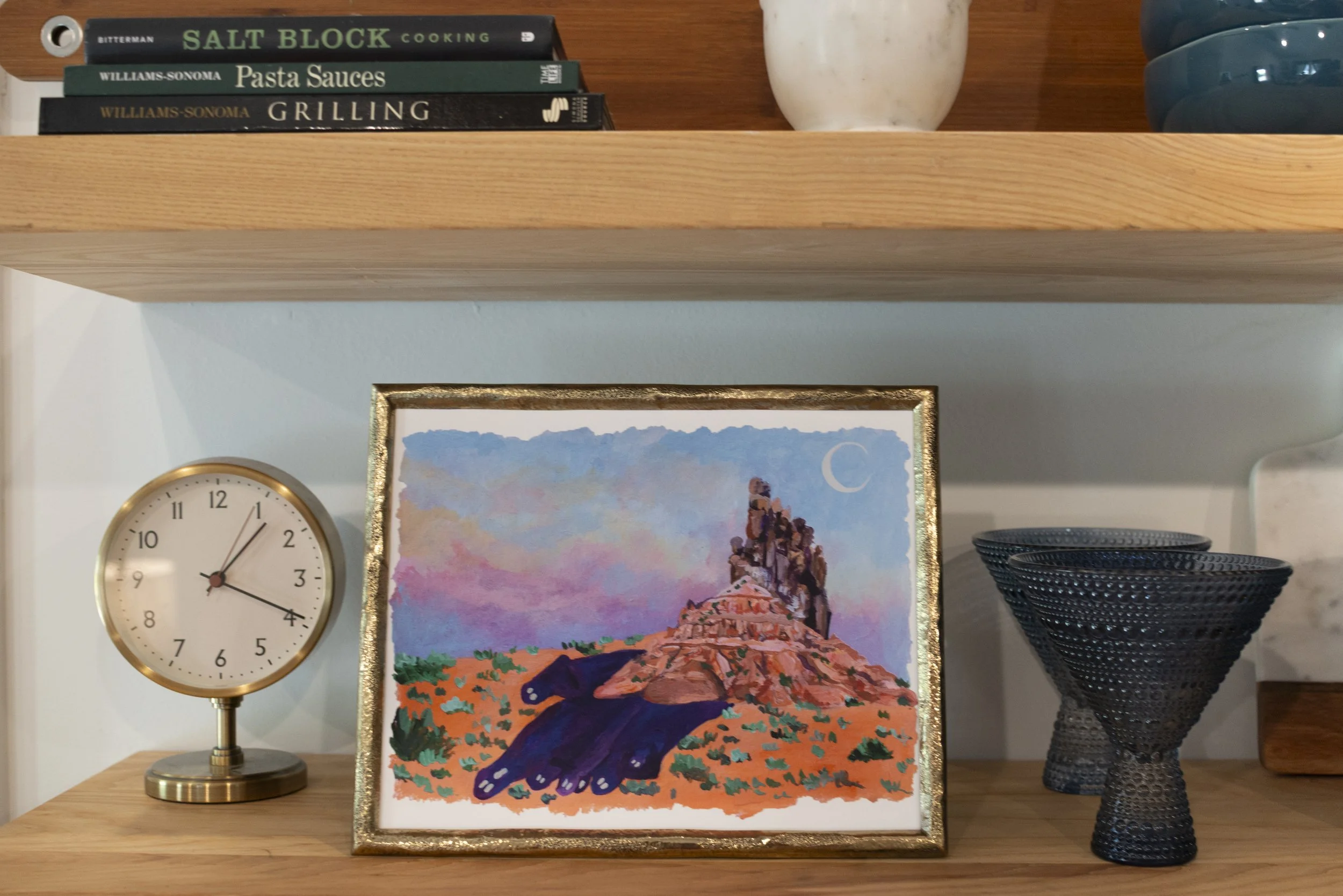 A print of a desert scene in a gold frame displayed on a wooden shelf with varioius kitchen glassware, a decorative gold clock, and cookbooks.