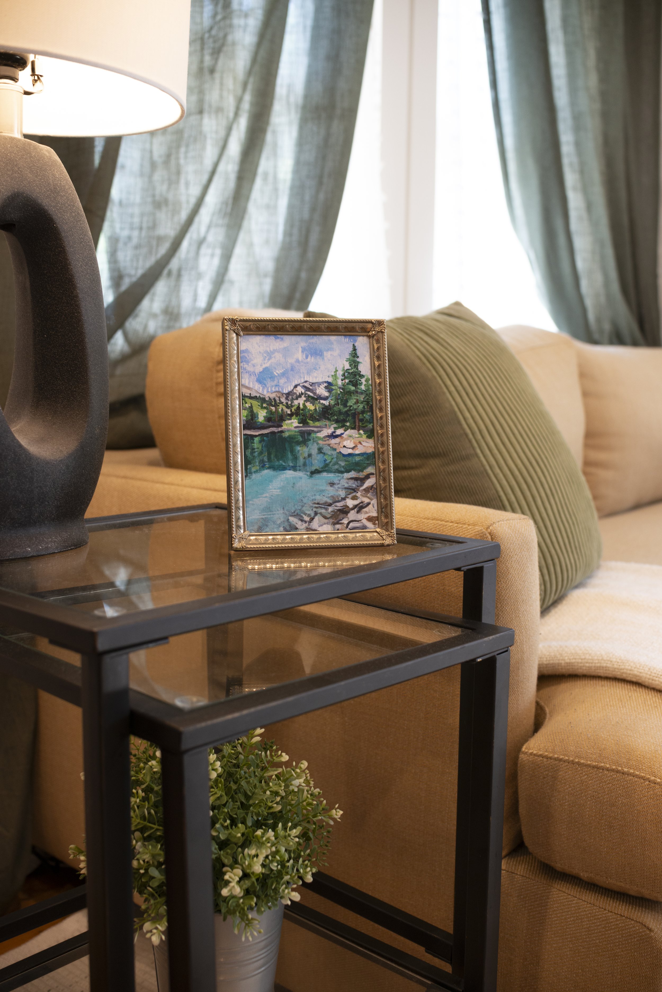Framed print of an alpine lake on a glass coffee table, styled with a lamp and indoor plant, set in front of a cozy living room couch.