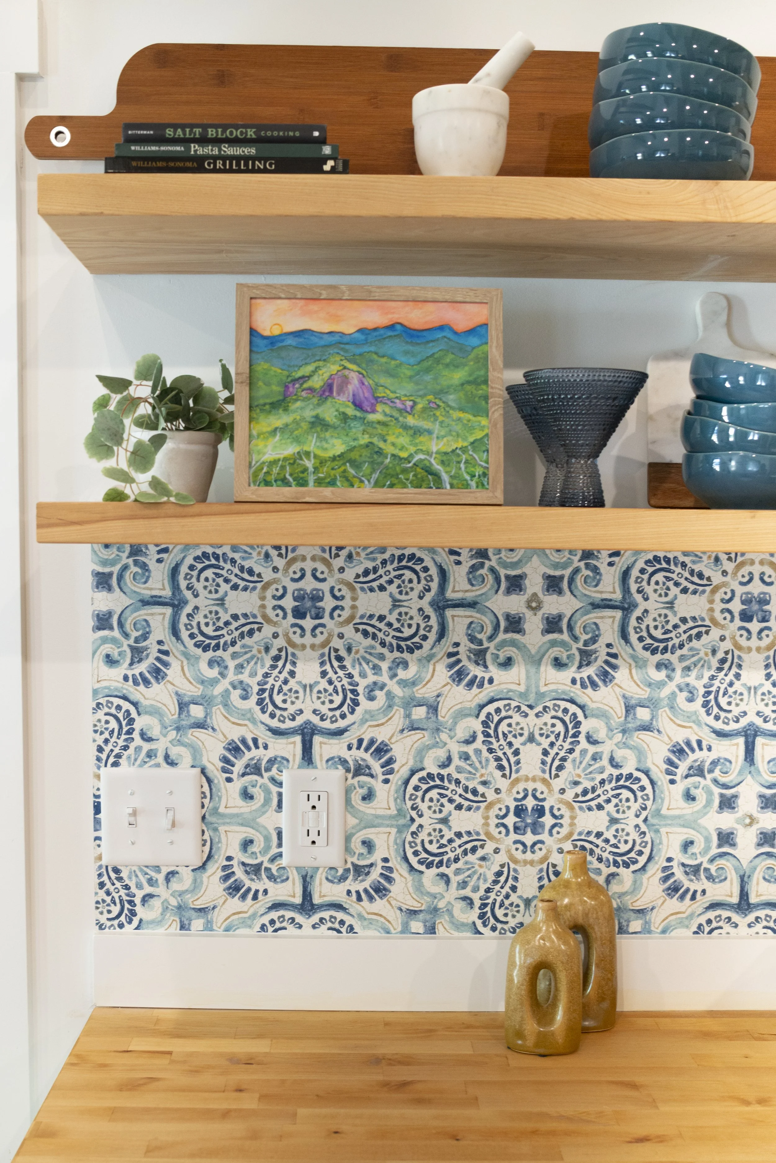 A framed print of a rock monolith surrounded by mountains, displayed on a kitchen shelf in a cozy, modern kitchen with wooden shelves and colorful accents.