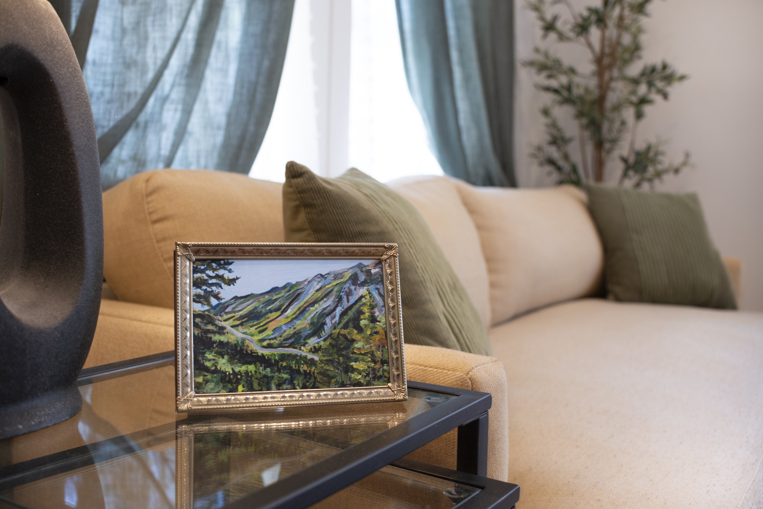 A mountain landscape print in a gold frame, displayed on a side table in a living room next to a cozy couch.