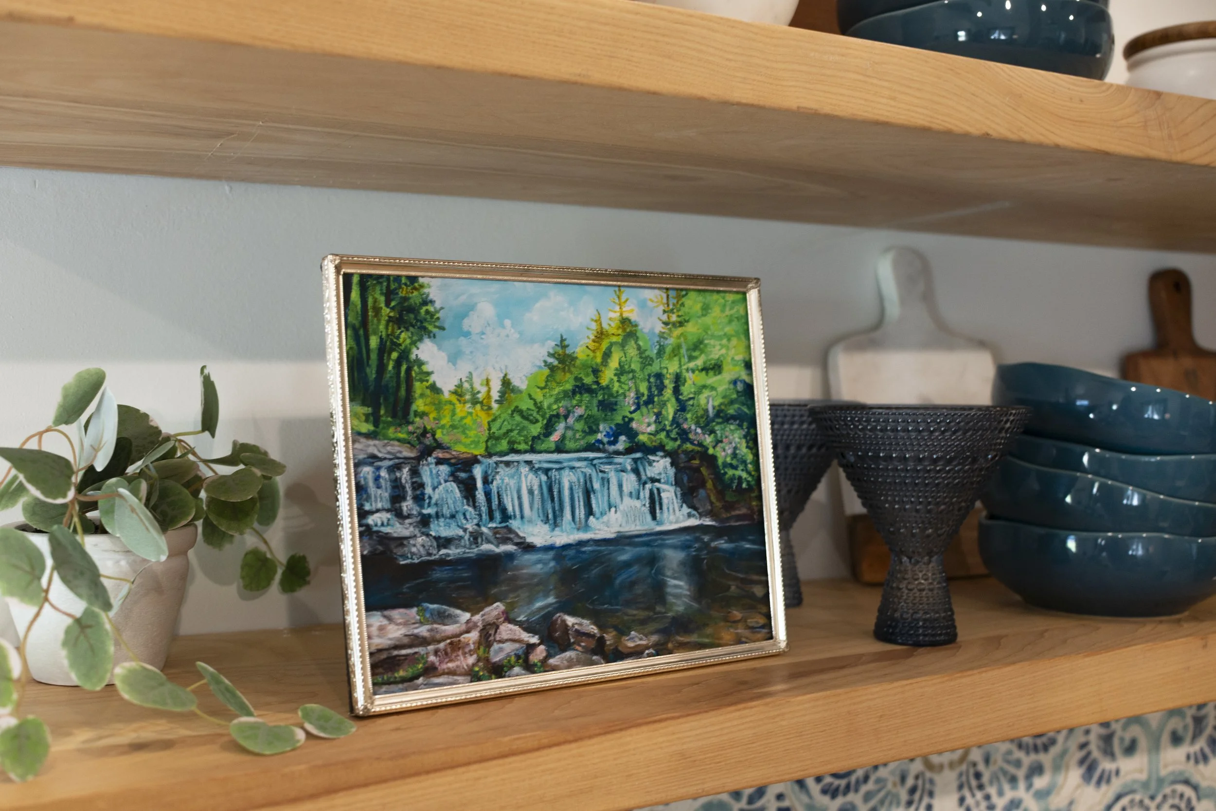 A side view of a framed print of Hooker waterfall in DuPont displayed on a wooden shelf with decorative dish ware, and an indoor plant.