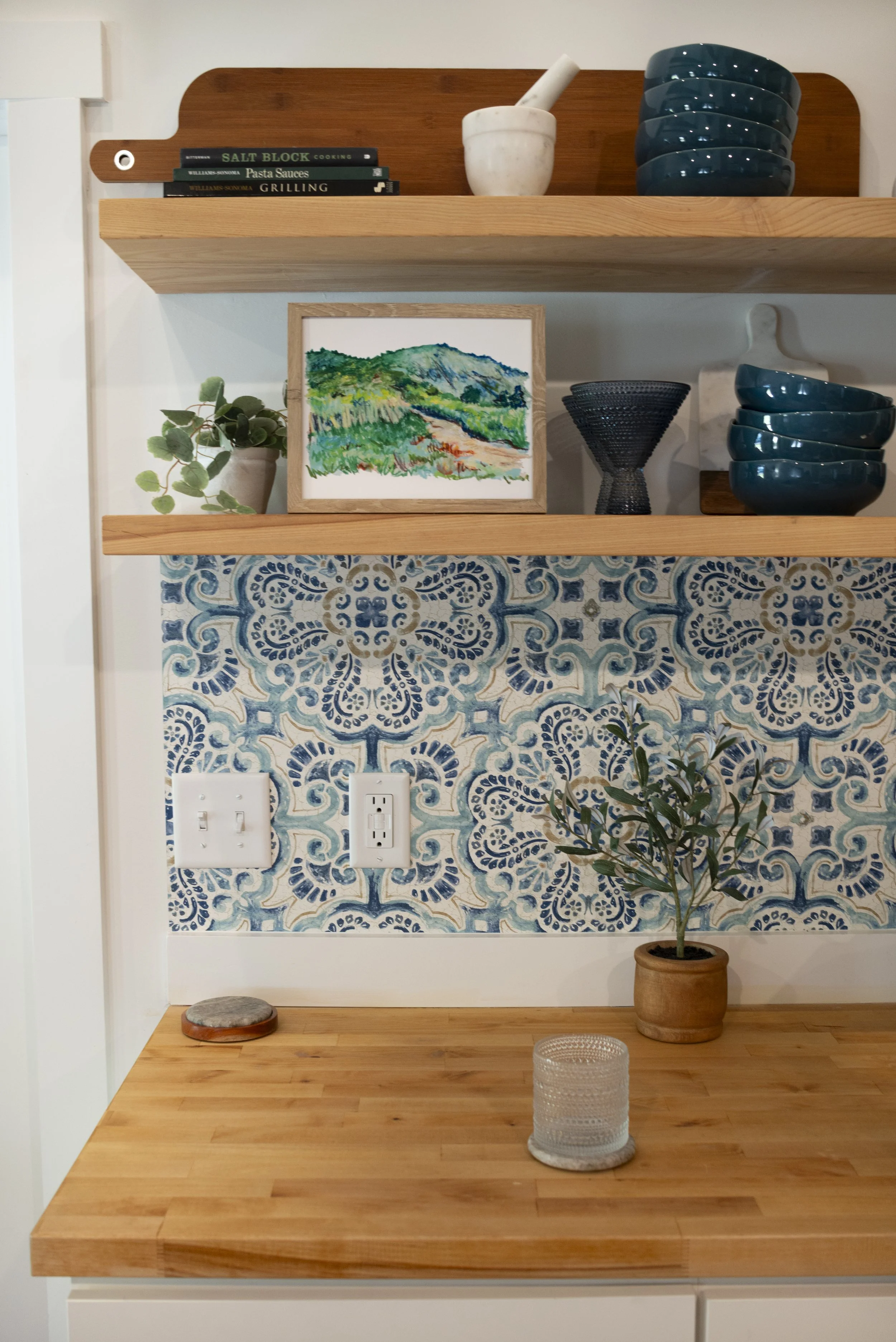 A framed print of an oil pastel painting of Black Balsam Knob displayed on a kichen shelf with decorative blue dish ware, indoor plants, and books.