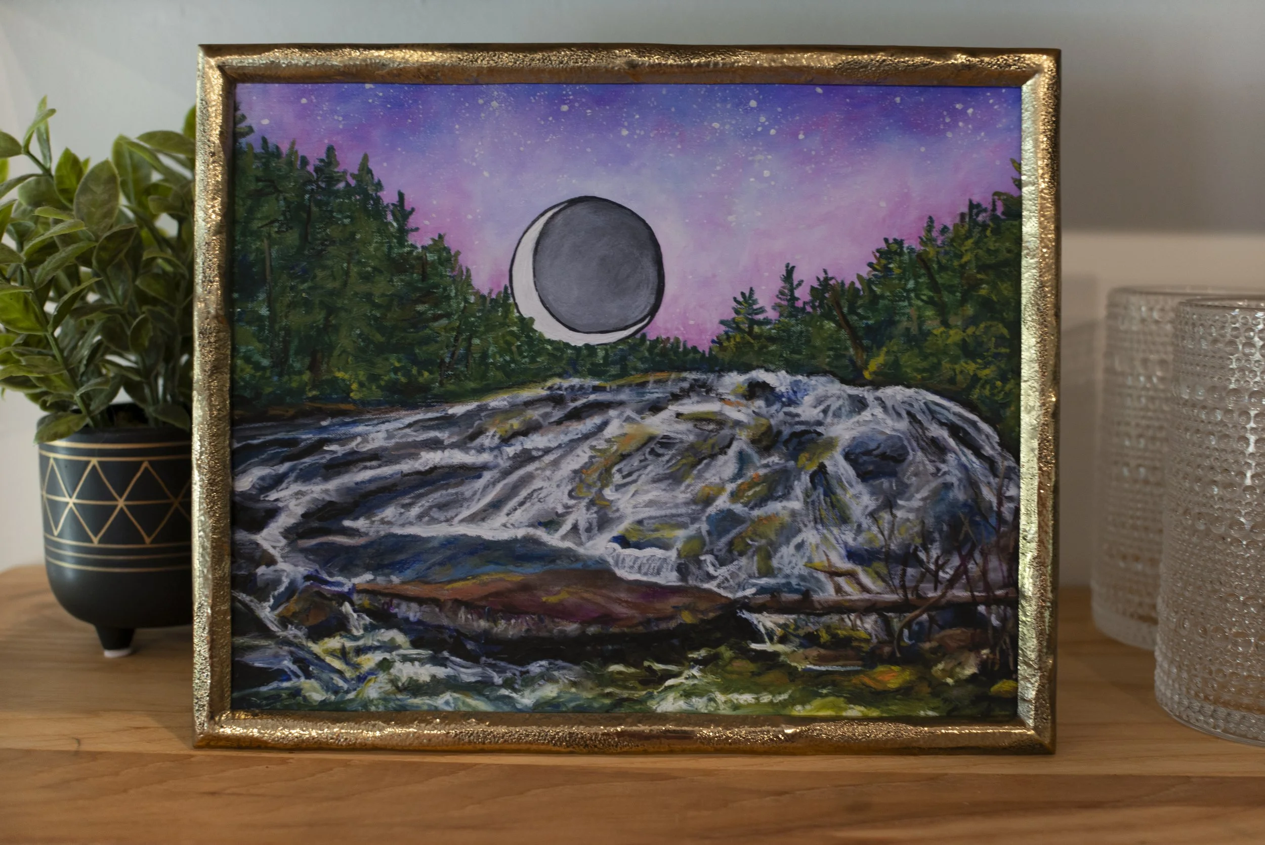 A print of Bridal Veil Falls in DuPont Forest in a gold frame between glassware and an indoor plant on a wooden shelf.