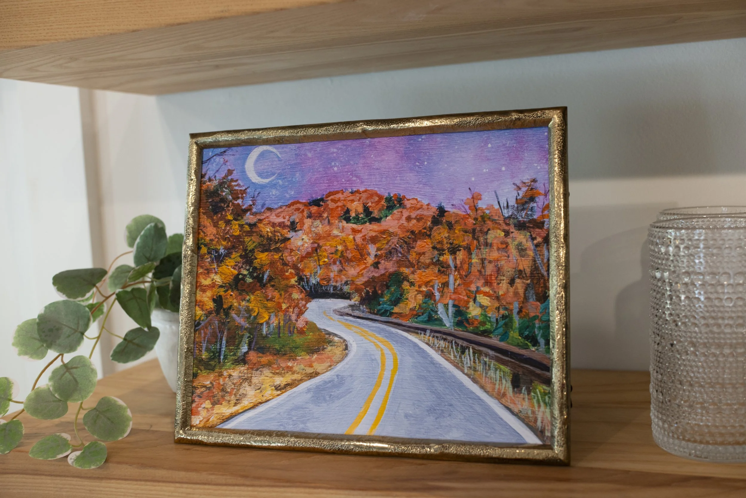 A framed print of the Blue Ridge Parkway in the fall displayed on a wooden shelf with an indoor plant and stylish glassware.