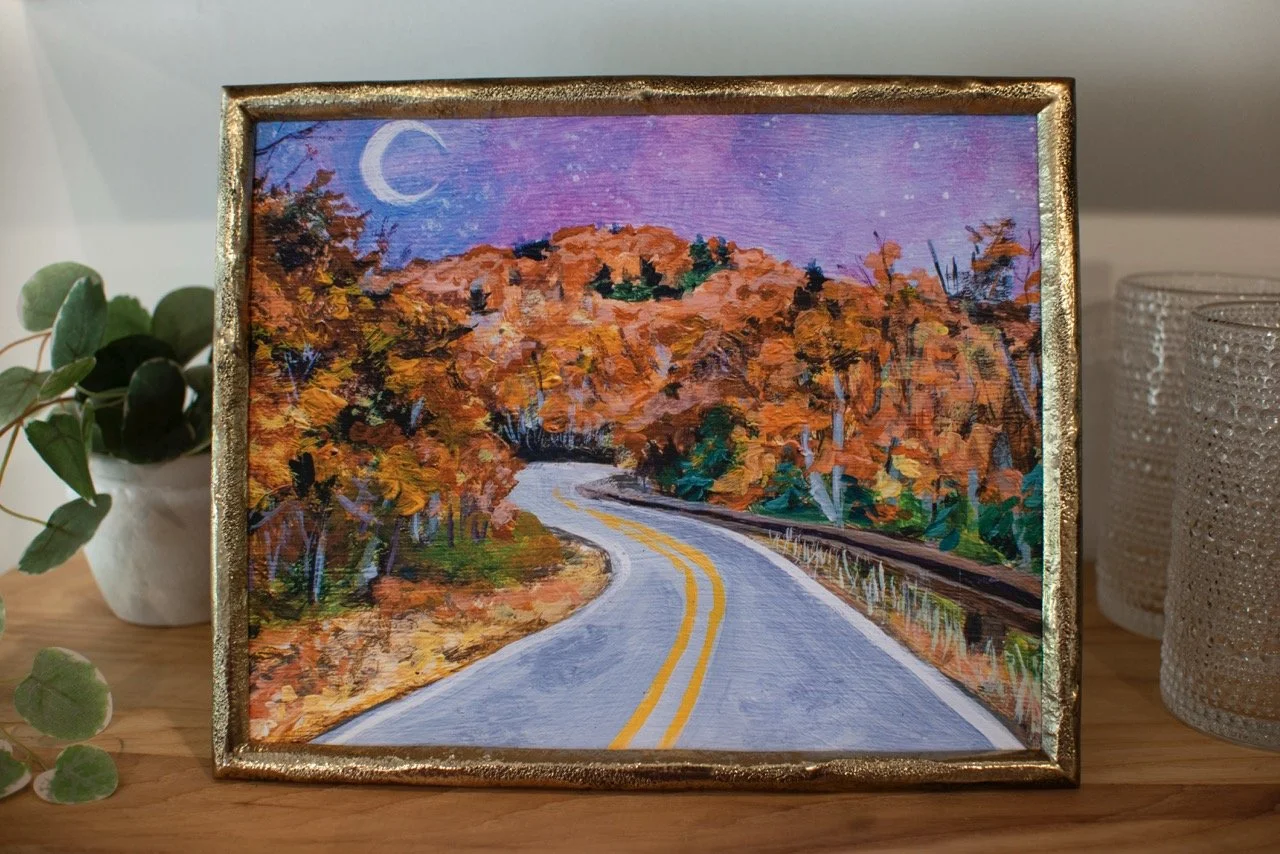 An autumn Blue Ridge Parkway art print displayed in a gold frame on a wooden shelf.