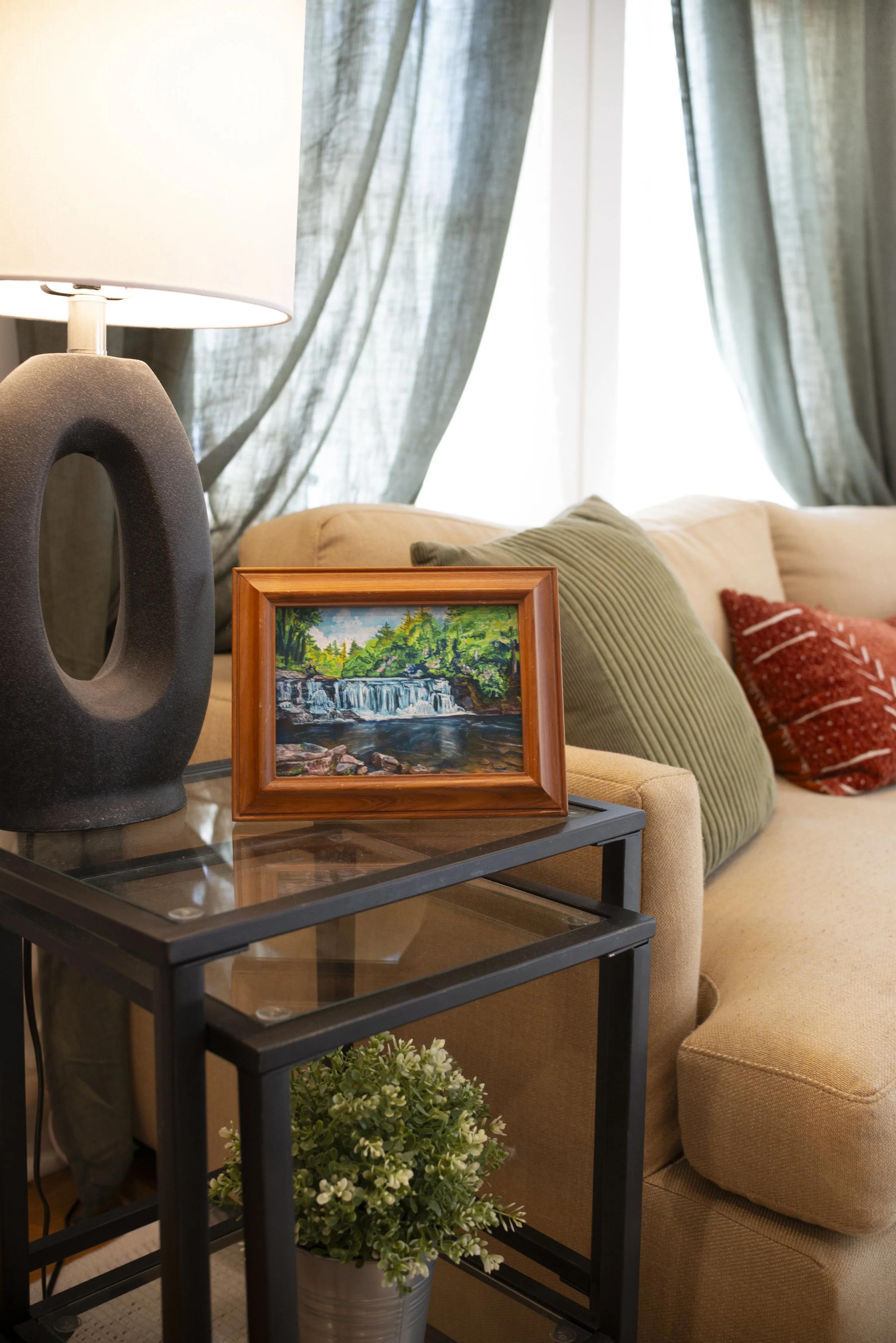 A fine art print of a DuPont waterfall in a gold frame, displayed on a side table in a modern living room next to a cozy couch.