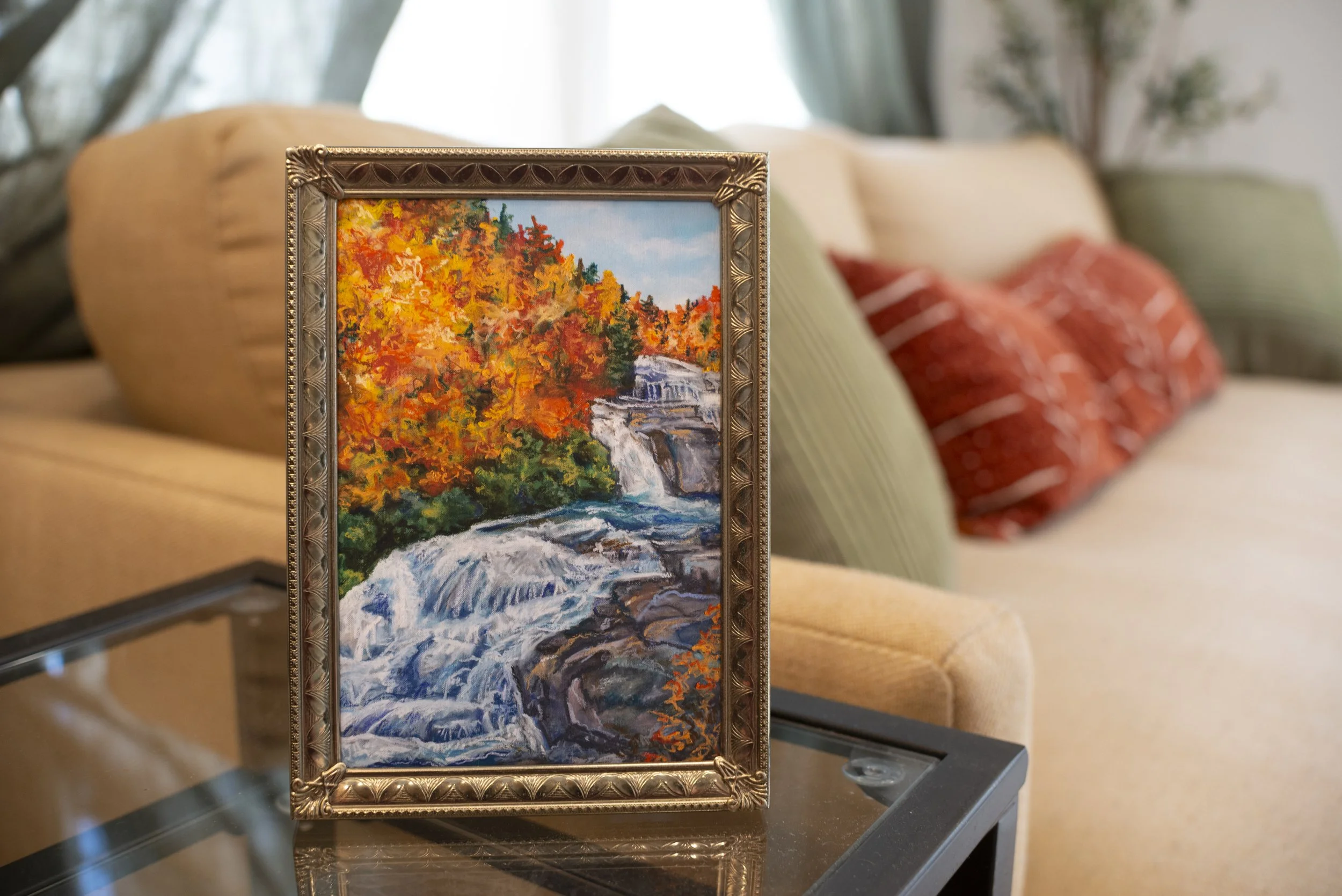 A print of a DuPont waterfall in the fall in a gold frame, displayed on a side table in front of a modern living room couch.