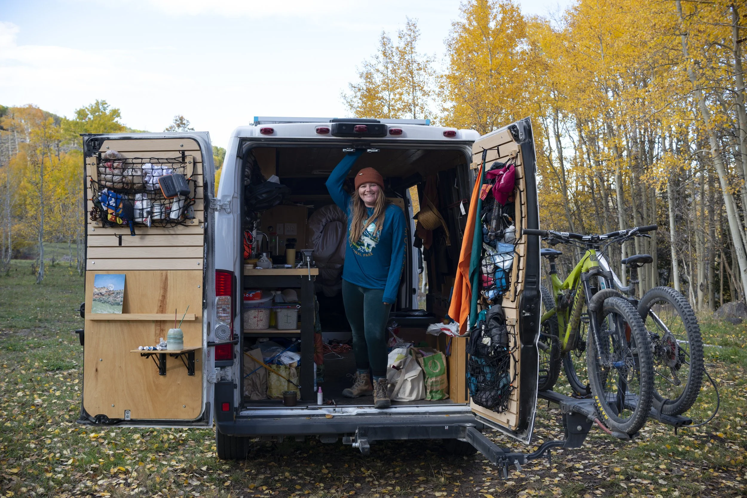 Artist standing in the open rear of a camper van, doors wide, against a backdrop of vibrant autumn foliage in an outdoor setting.