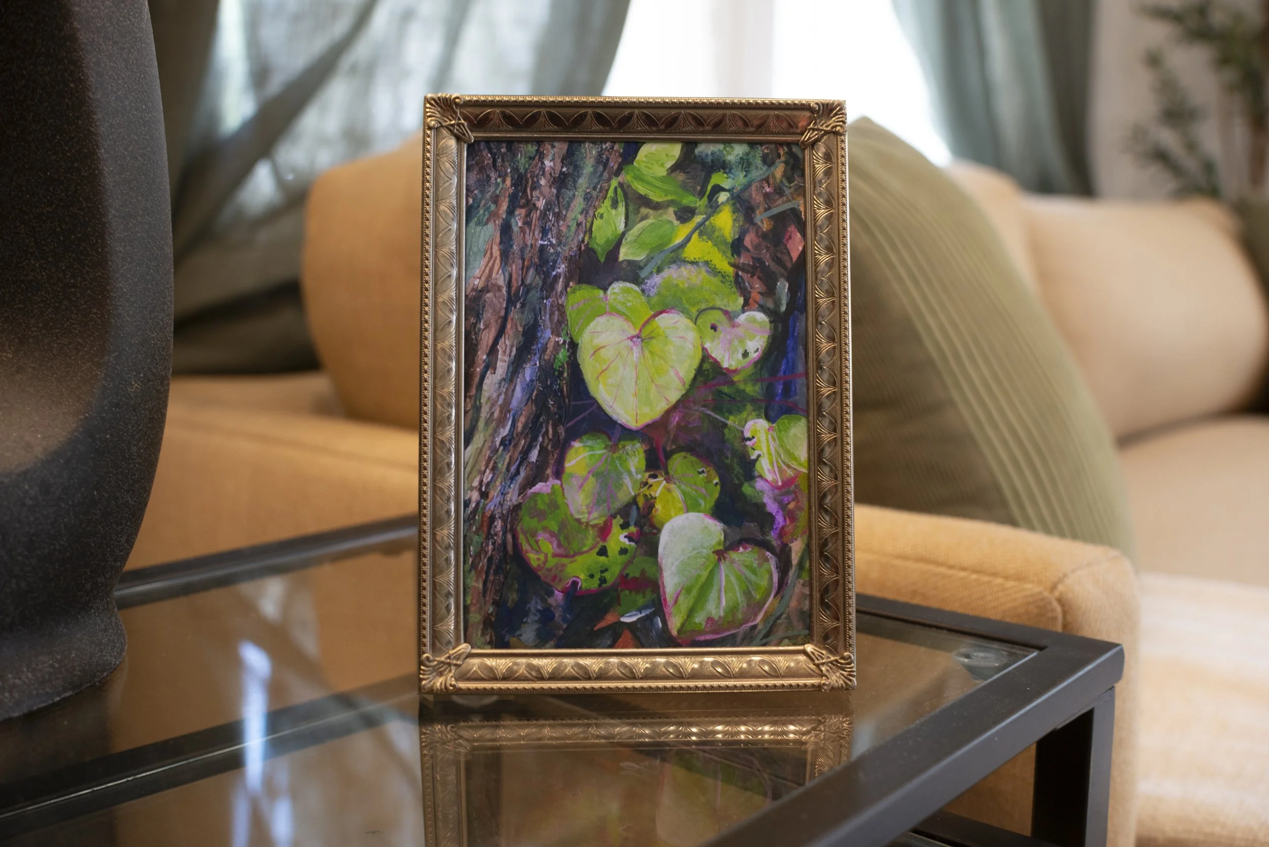 A botanical fine art print in a gold frame, on a glass side table in front of a beige couch with a green pillow.
