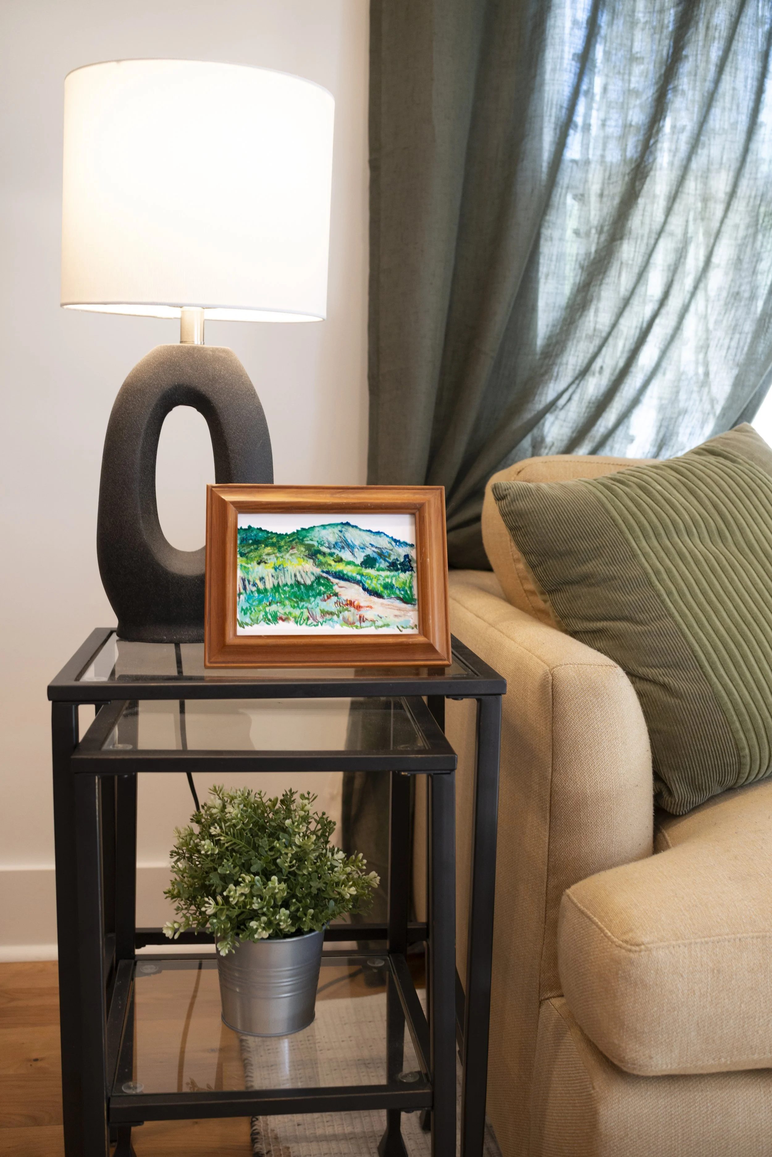 An Appalachian mountain landscape print in a wooden frame, displayed on a side table with a lamp and houseplant in a living room next to a cozy couch.