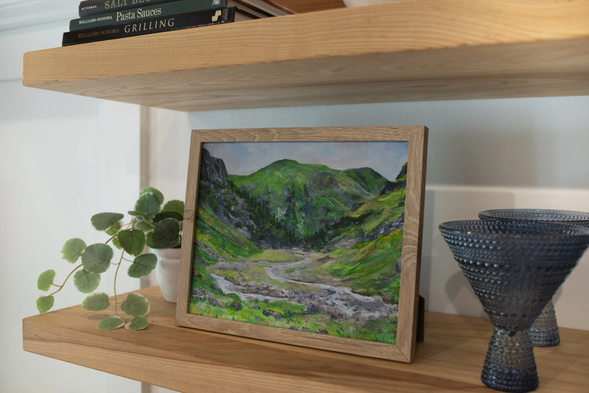 A side view of a print of Glencoe in the Scottish Highlands in a wooden frame displayed on a cozy shelf with decorative glasses and an indoor plant.