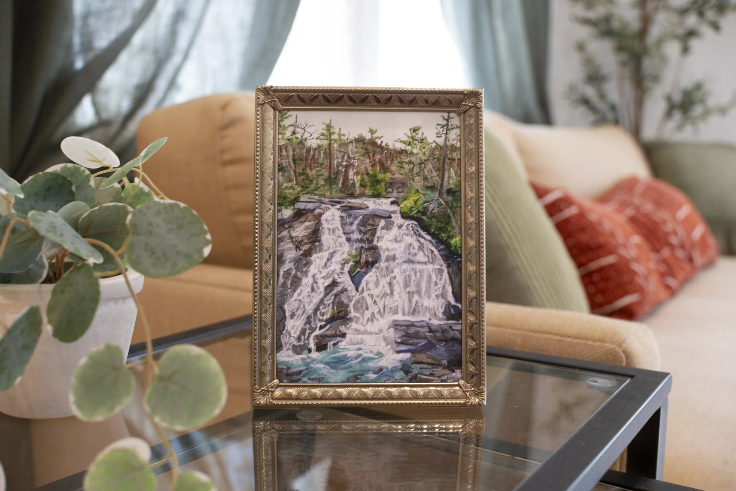 A print of a DuPont waterfall in a gold frame, displayed on a side table in a living room next to a cozy couch and indoor plant.