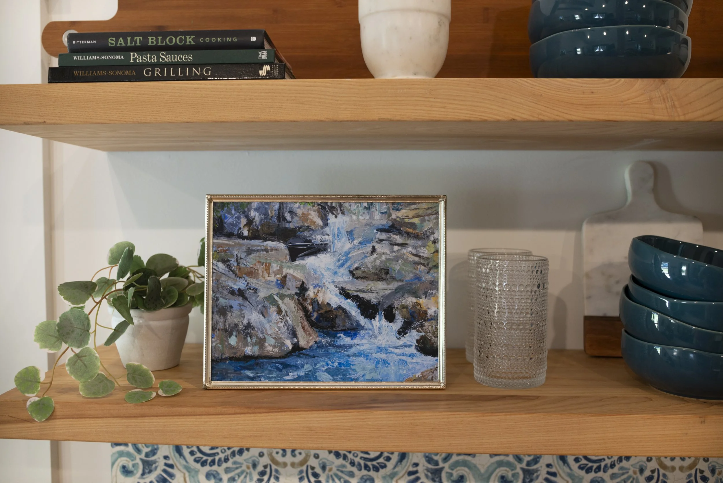 A print of a Western NC waterfall in a gold frame displayed on a shelf with glasses, dish ware, an indoor plant, and cookbooks, viewed from the side.