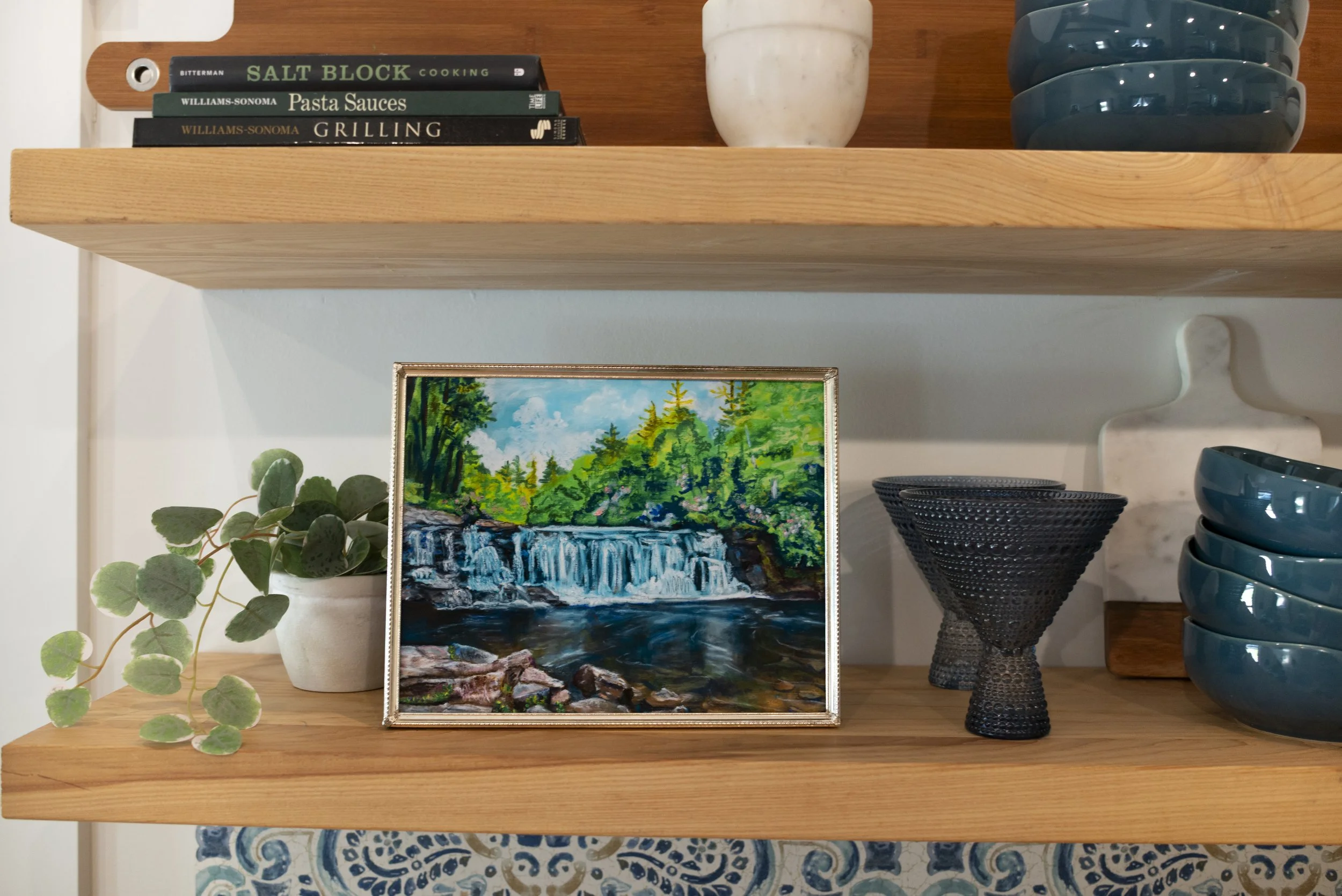 A framed print of Hooker waterfall in DuPont displayed on a wooden shelf with decorative dish ware, cookbooks, and an indoor plant.