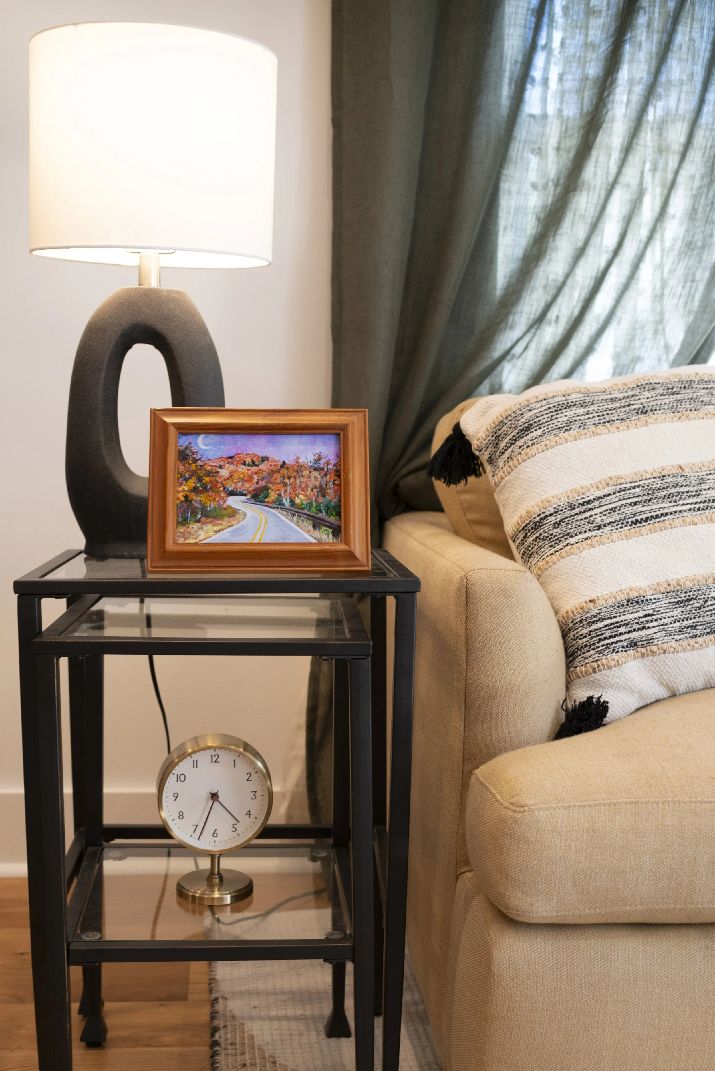 A framed print of a Blue Ridge Parkway overlook in the fall, displayed on a side table in a modern living room with a cozy lamp, next to a couch.