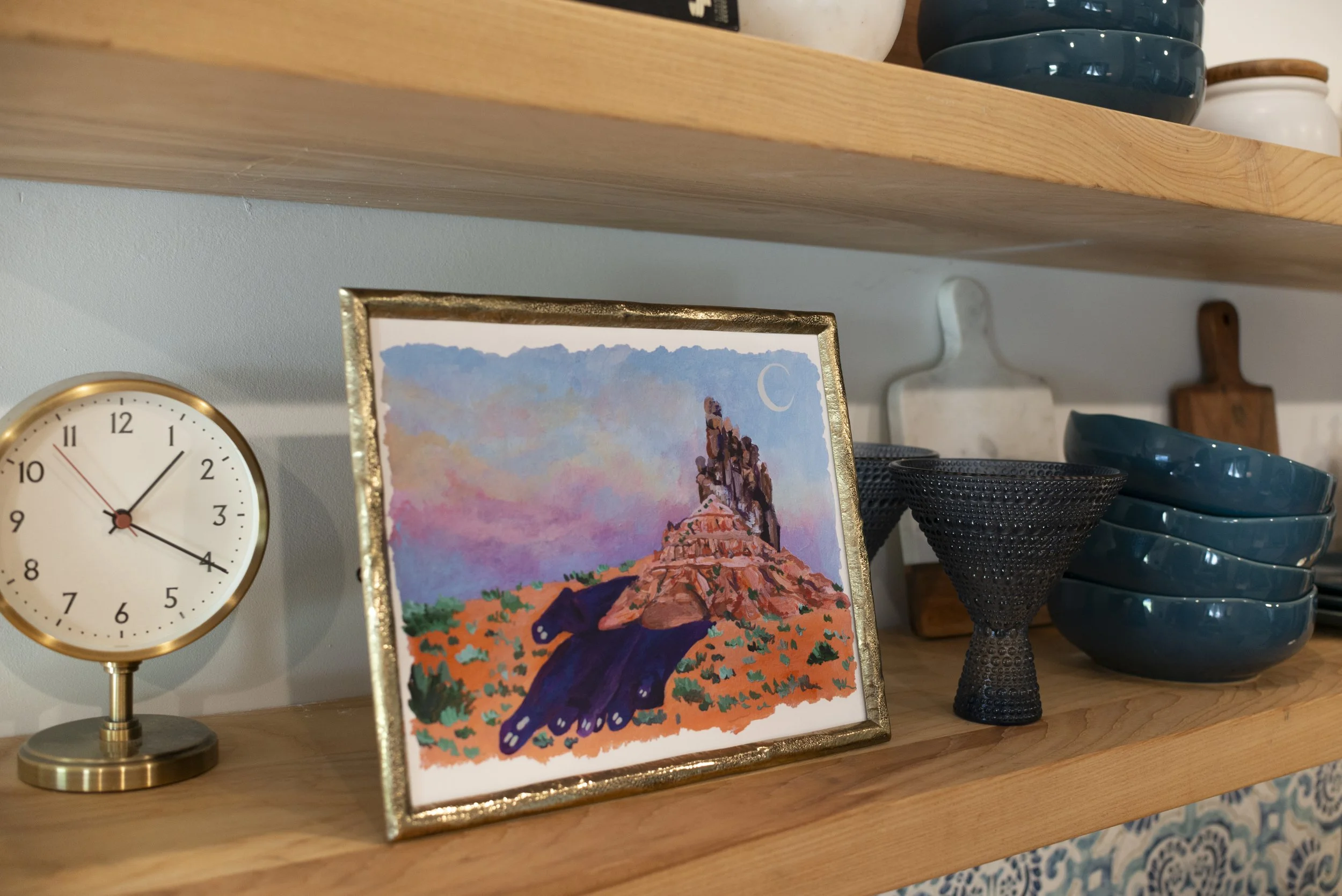 A side view of a desert print in a gold frame on a wooden shelf with kitchen glassware, a gold clock, and cookbooks, adding style to the space.