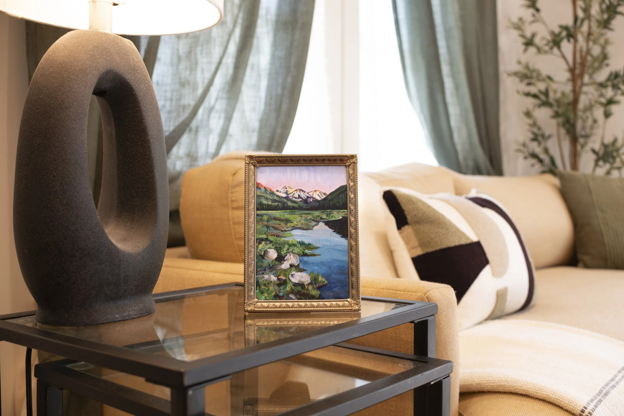 A print of a mountain landscape in a gold frame displayed in a modern living room on a glass living room table next to a lamp and in front of a couch.