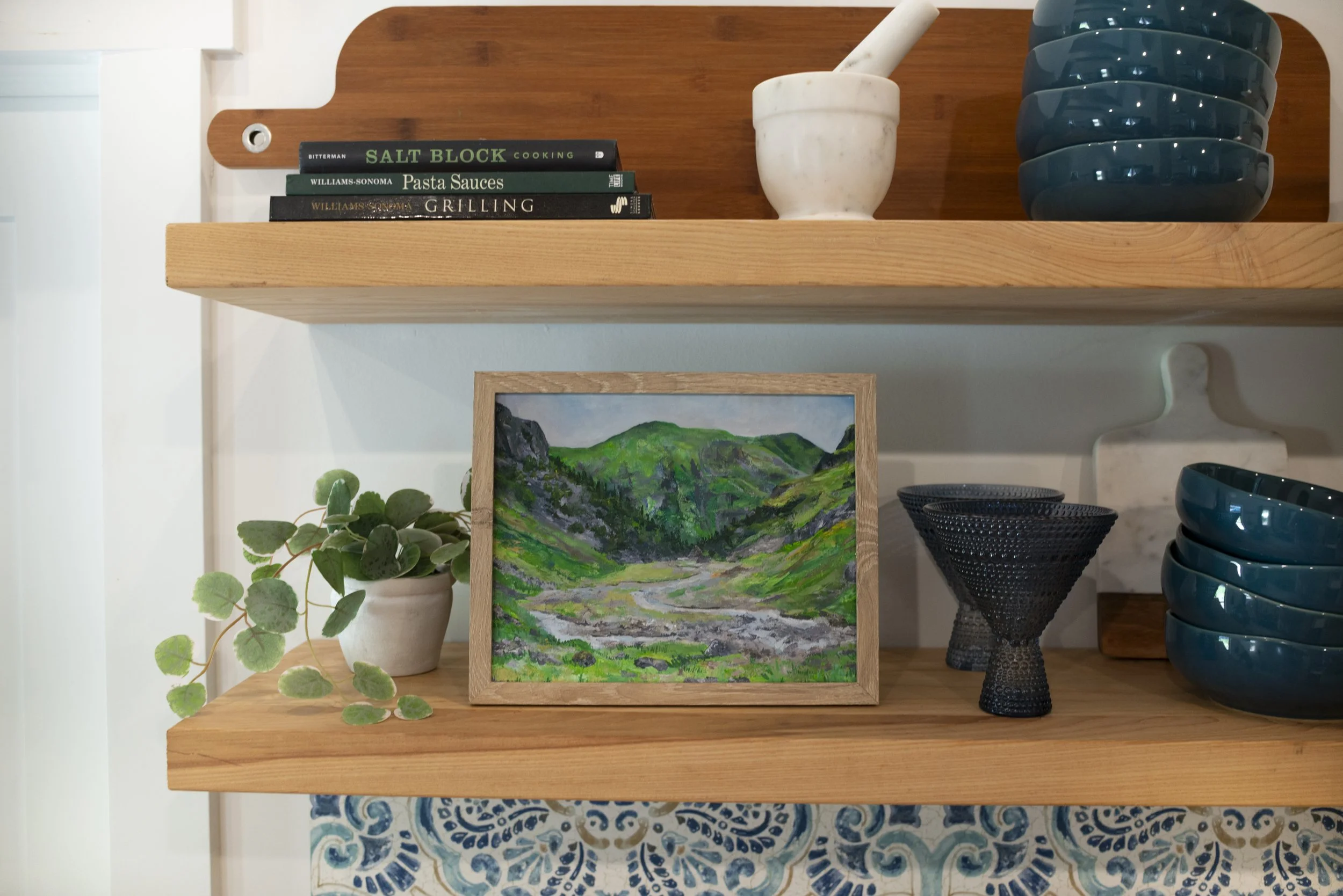 A print of Glencoe in the Scottish Highlands in a wooden frame displayed on a kichen shelf with decorative blue dish ware, an indoor plant, and books.