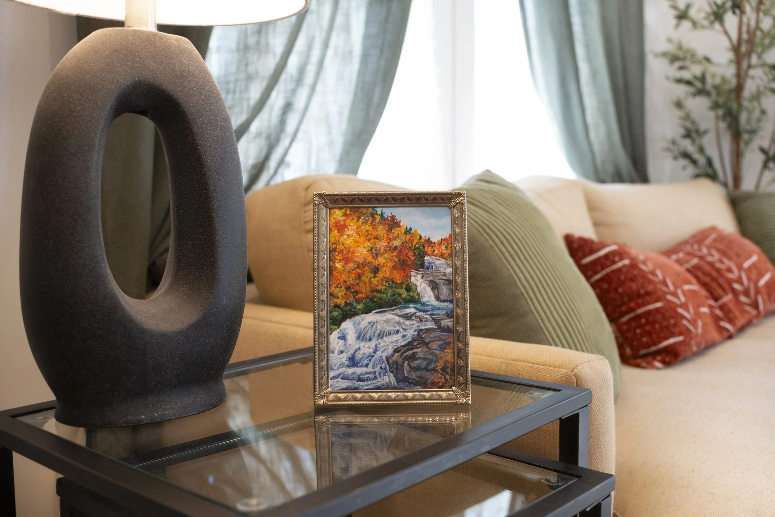 A framed print of a DuPont waterfall in the fall, displayed on a side table in a modern living room next to a couch.