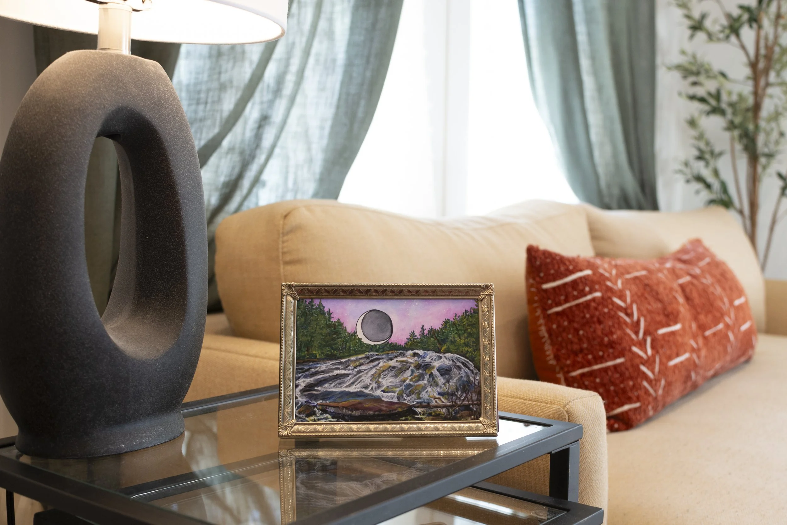 A print of Bridal Veil Falls in DuPont Forest in a gold frame displayed on a glass living room table next to a lamp and in front of a couch.