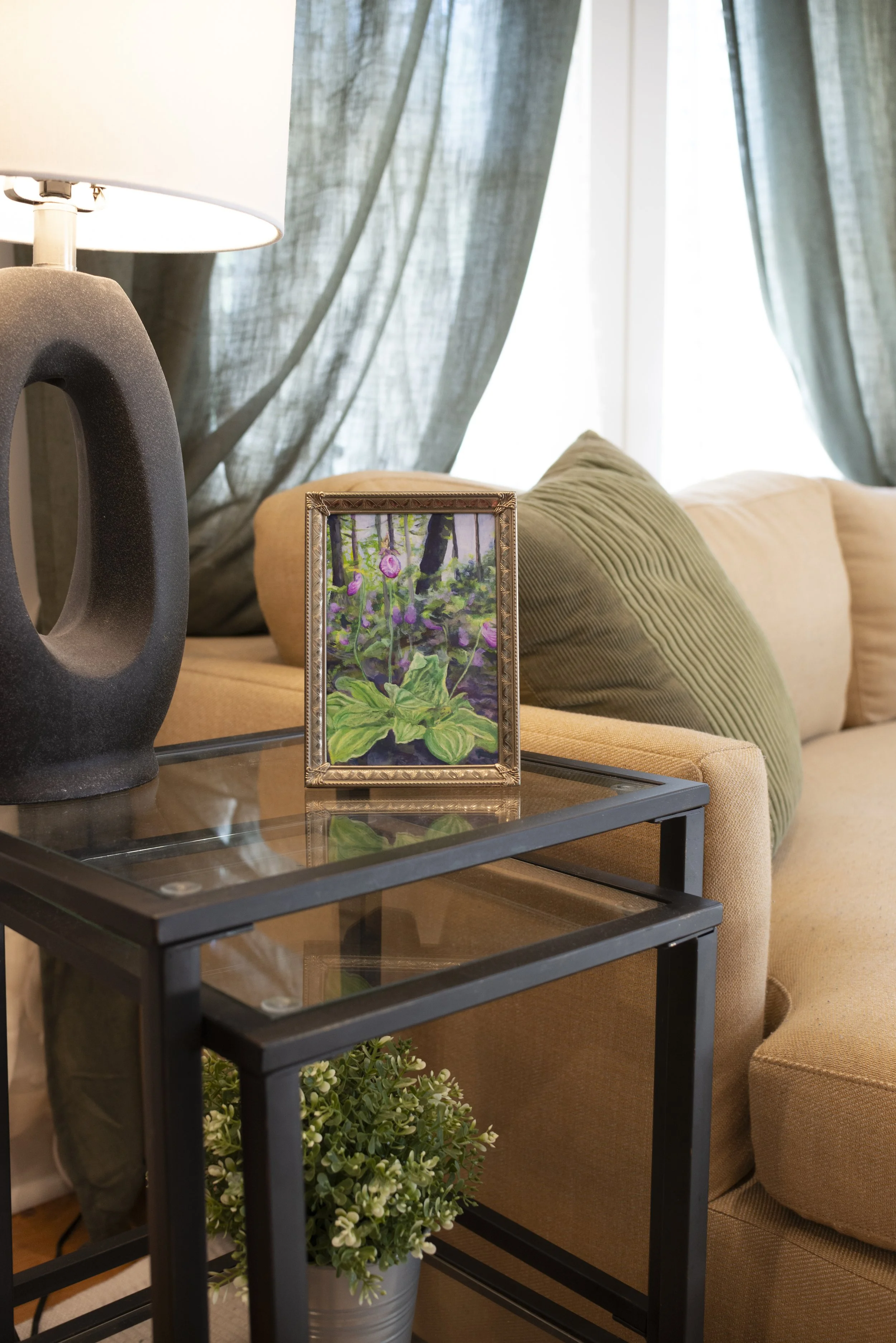 A fine art print of pink Lady Slipper Orchids in a gold frame, displayed on a side table next to a cozy couch and indoor plant in a modern living room.