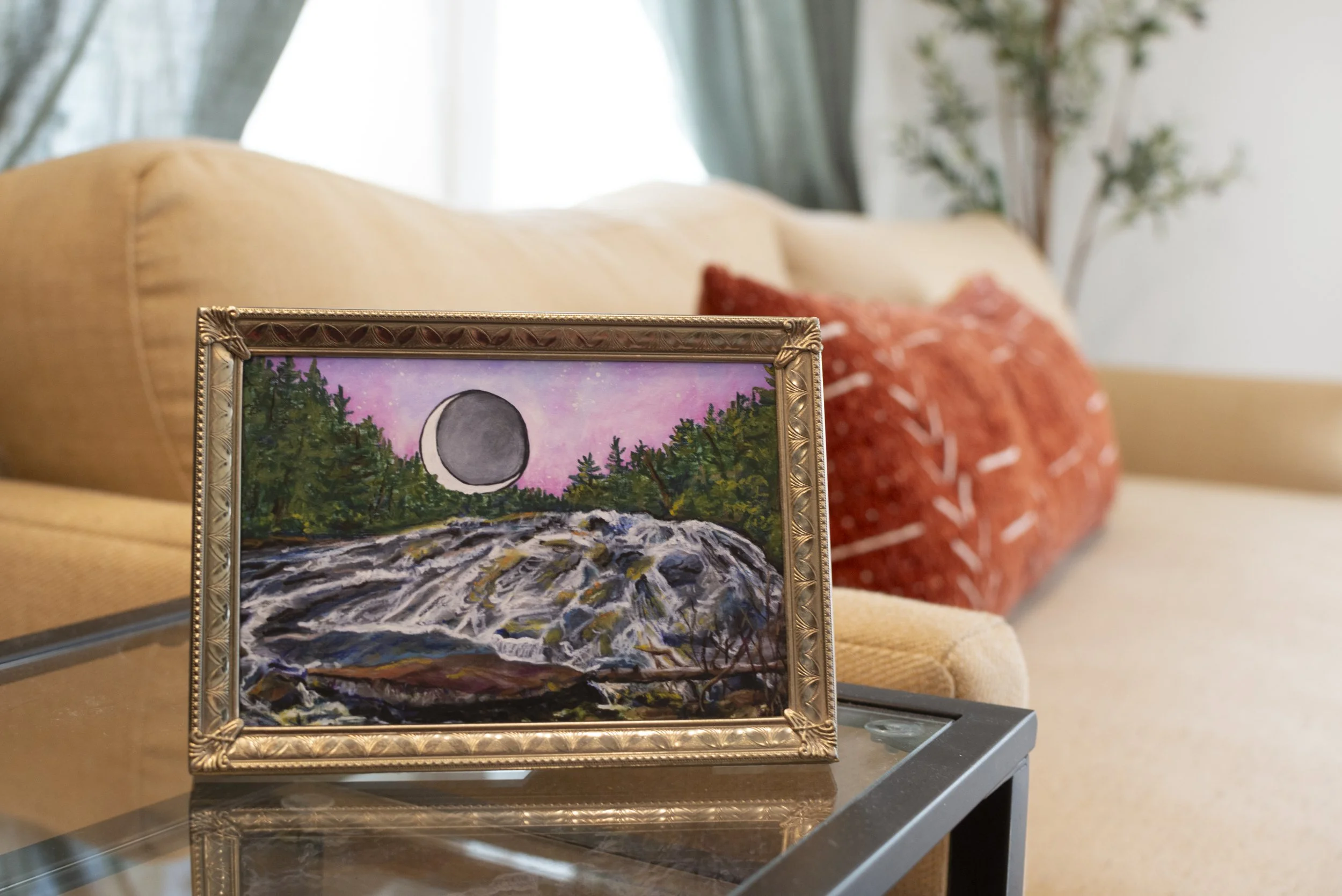 A print of Bridal Veil Falls in DuPont Forest in a gold frame displayed on a glass living room table in front of an oversized couch.