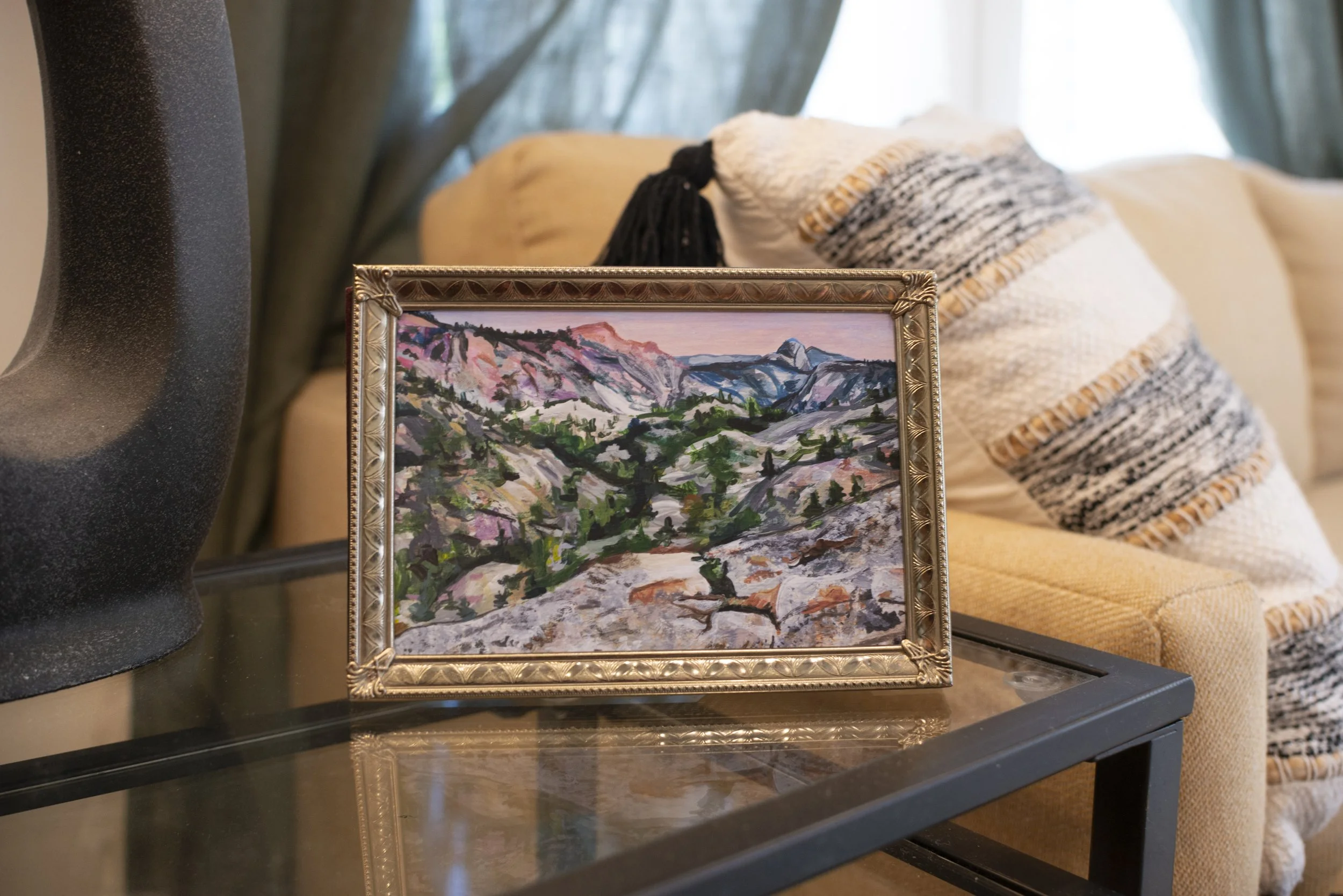 A Yosemite landscape print in a gold frame, displayed on a side table in a living room next to a cozy couch with a bohemian patterned pillow.