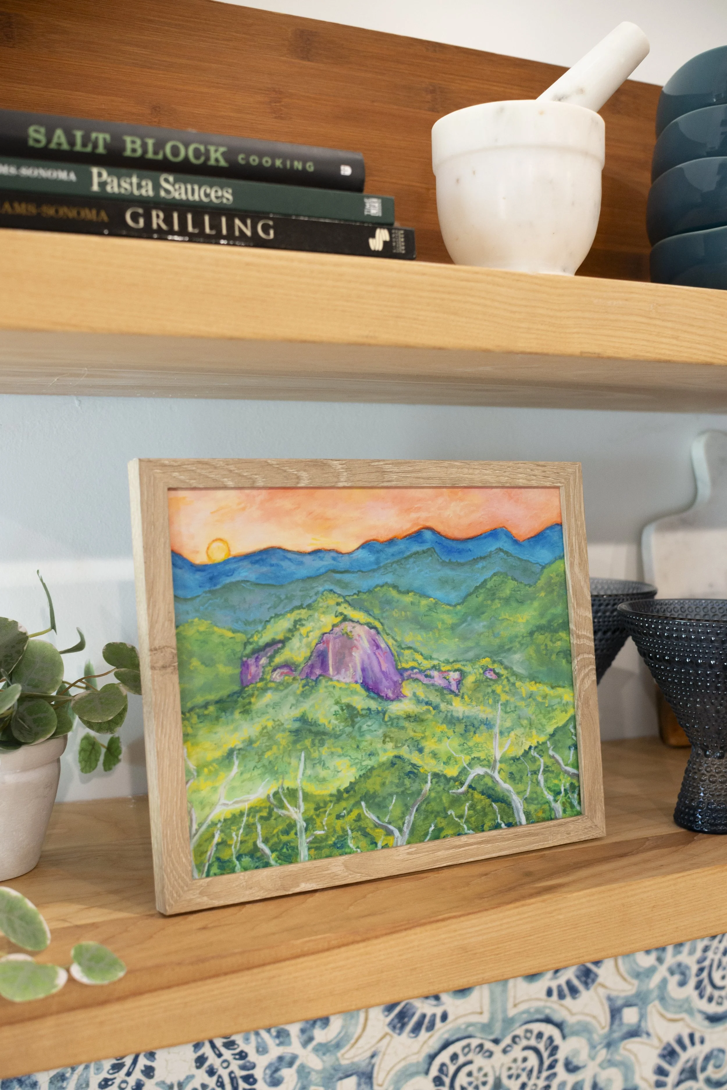 A framed print of a rock monolith surrounded by mountains displayed on a shelf with decorative vases, glasses, dish ware, and cookbooks, viewed from the side.