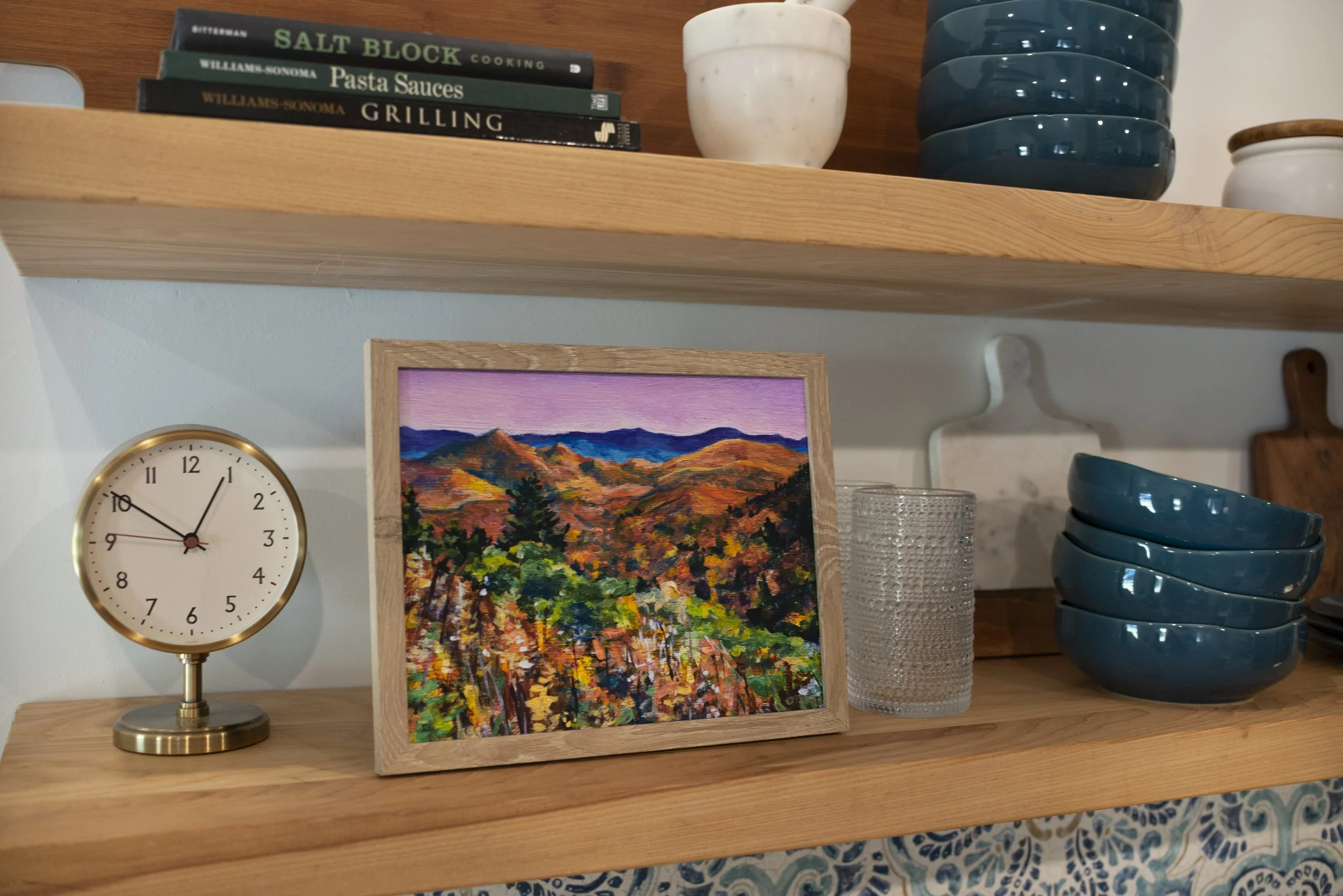 A framed print of a Blue Ridge Parkway overlook in the fall displayed on a shelf with a decorative clock, glasses, dish ware, and cookbooks.