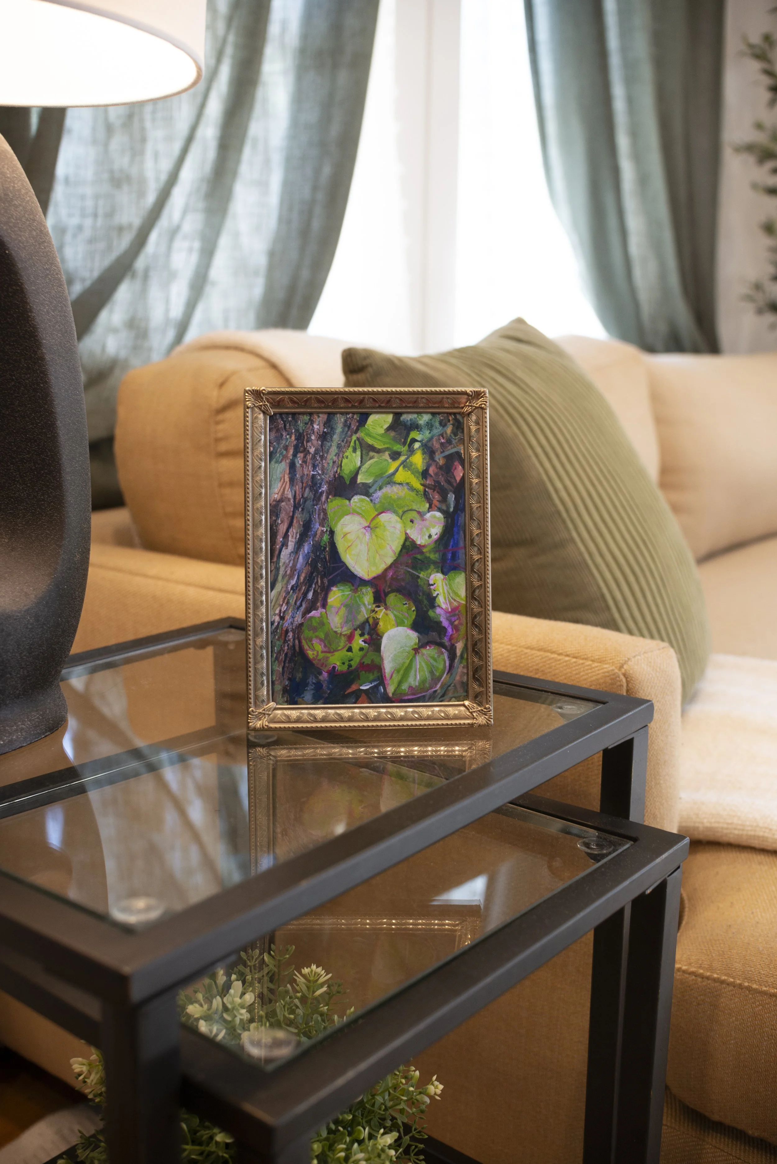 Between-The-LogA print of forest plant life in a gold frame, displayed on a side table next to a black lamp in a living room next to a couch with a green pillow.s-Table-Decor.jpg