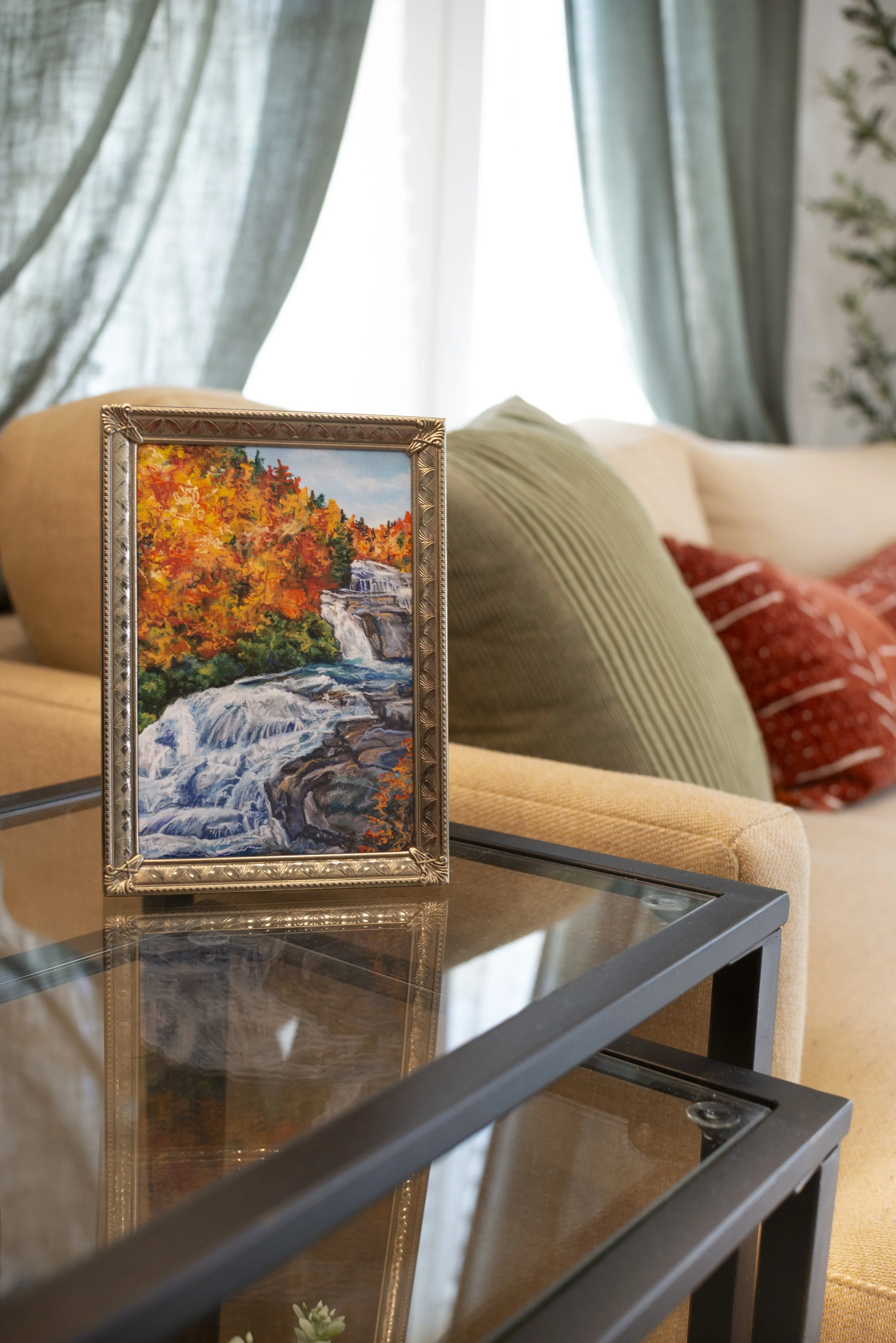 A framed print of a DuPont waterfall in the fall, displayed on a side table in a modern living room next to a couch sitting in front of a window.