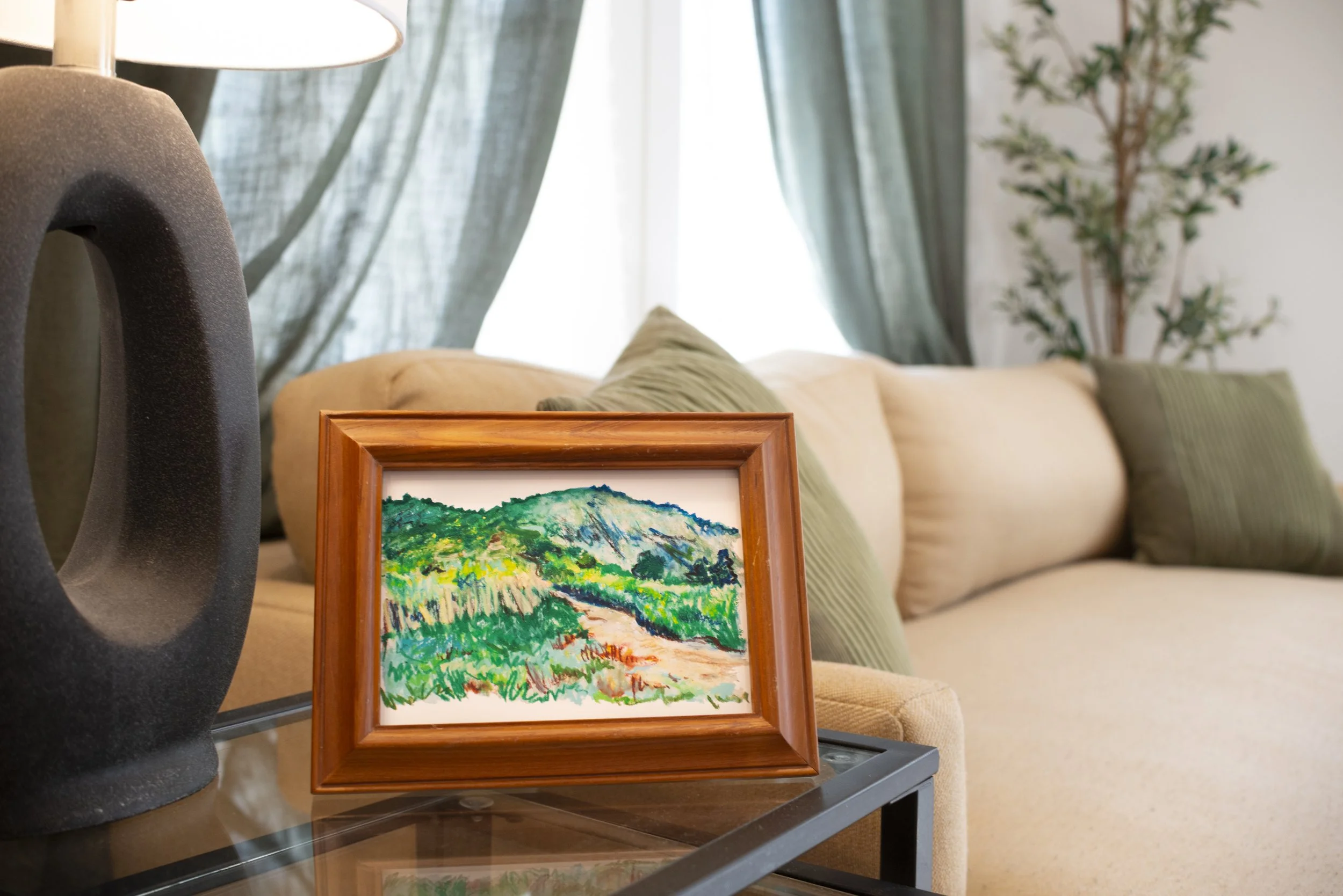 An Appalachian mountain landscape fine art print in a wooden frame, displayed on a glass side table next to a cozy couch with green pillows.