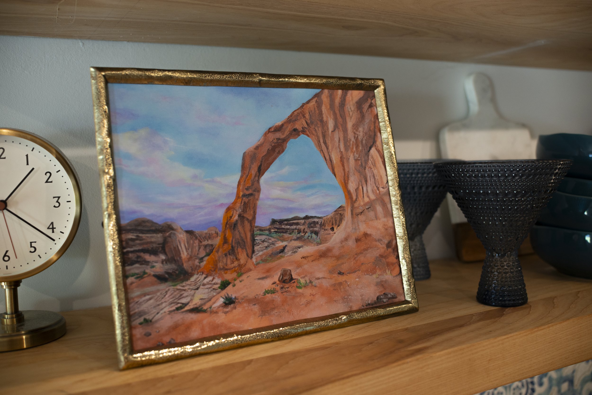 A side view of a print of Corona Arch in Moab, UT in a gold frame displayed on a cozy shelf with blue glassware and decorative gold clock.