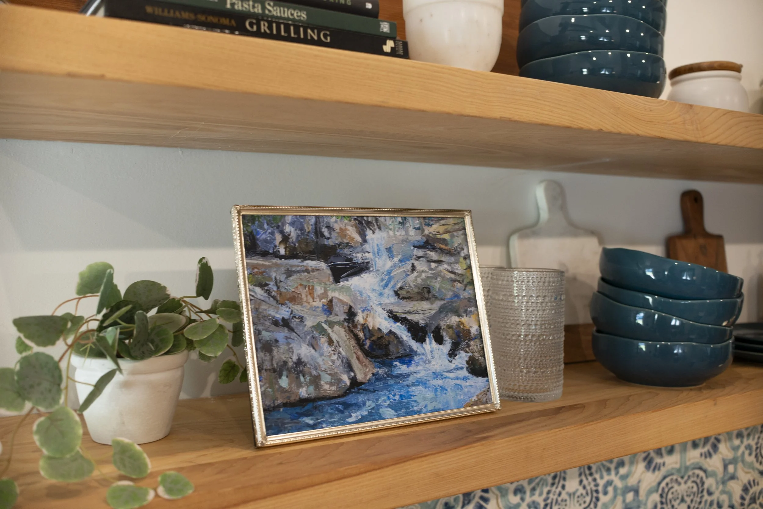 A print of a Western NC waterfall in a gold frame displayed on a shelf with glasses, dish ware, an indoor plant, and cookbooks.