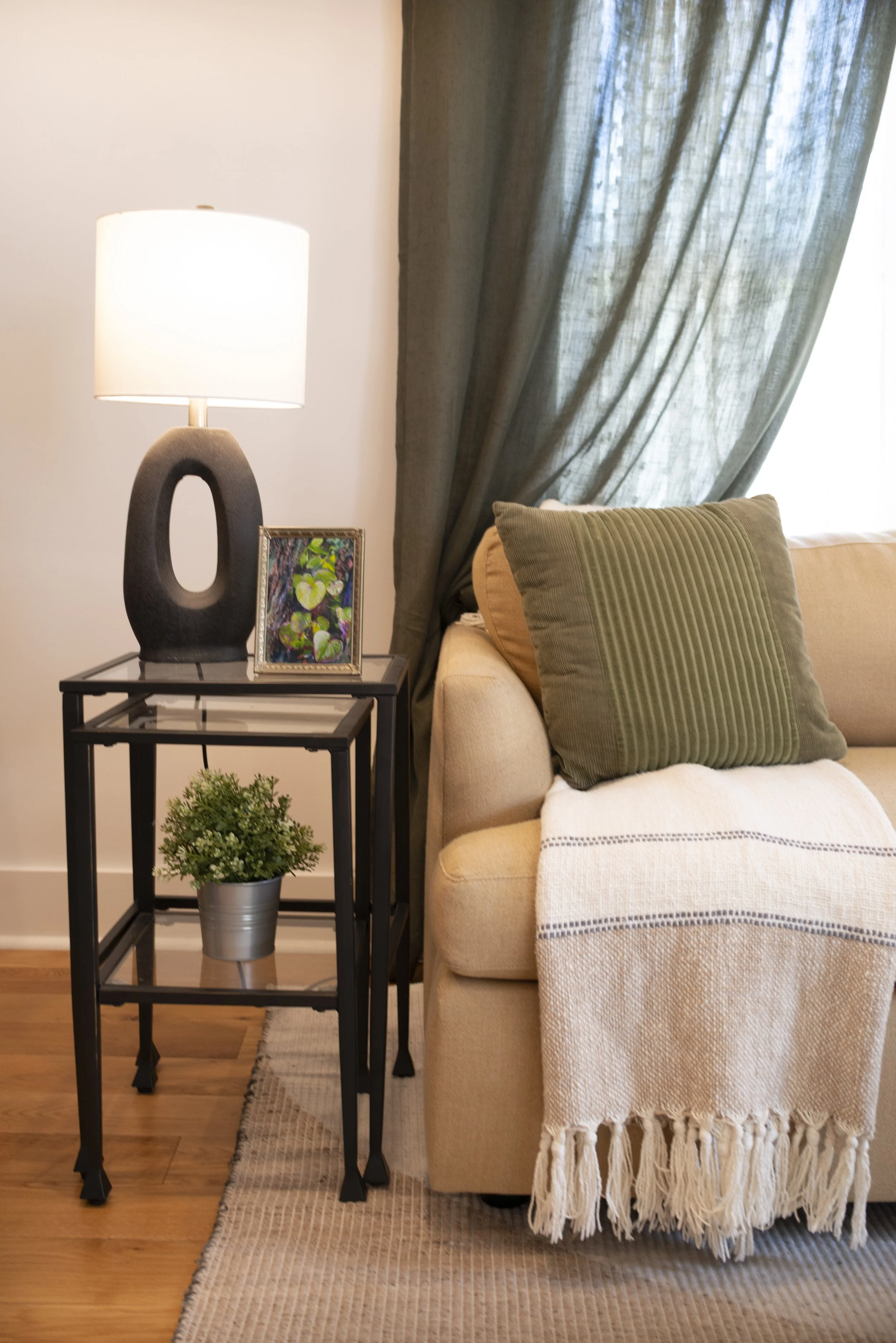 Framed forest plant art print on a glass side table with a modern lamp and potted plant beside a cozy couch with a green pillow and throw blanket.