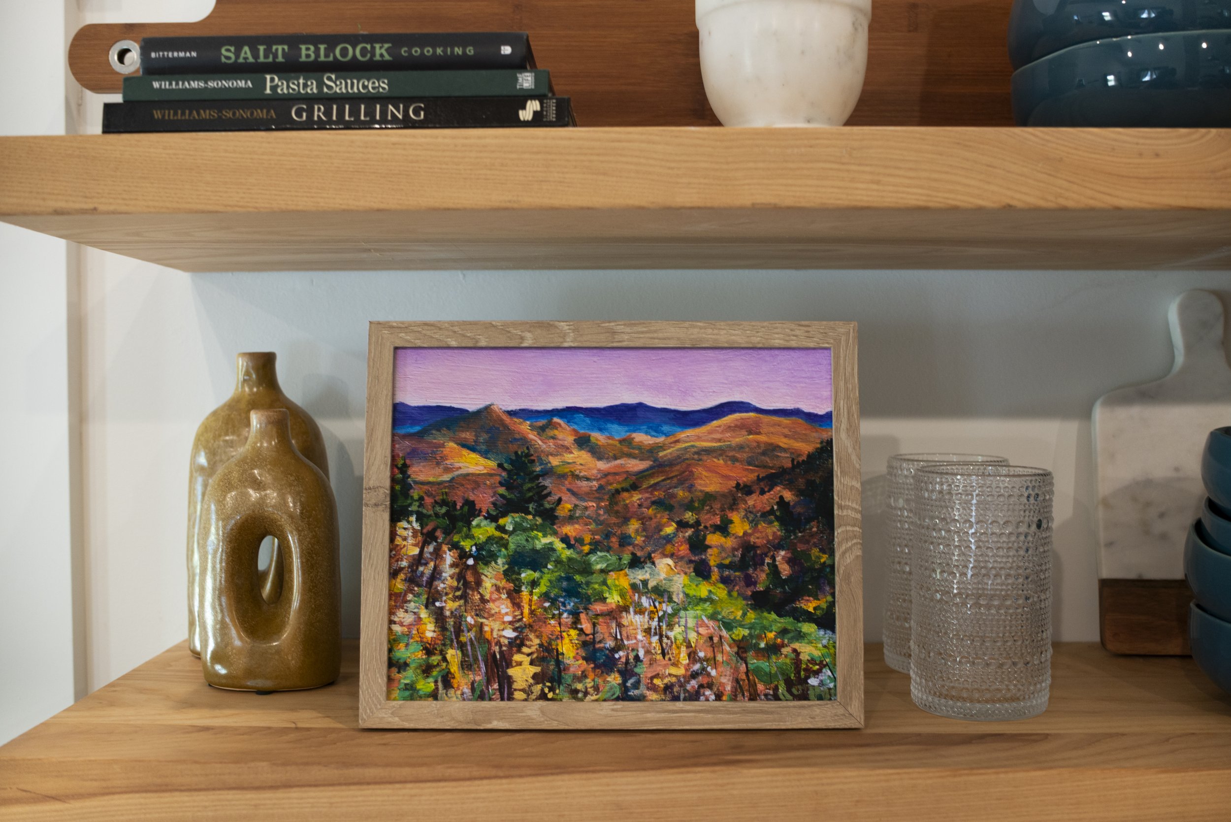 A framed print of a Blue Ridge Parkway overlook in the fall displayed on a shelf with decorative vases, glasses, dish ware, and cookbooks.