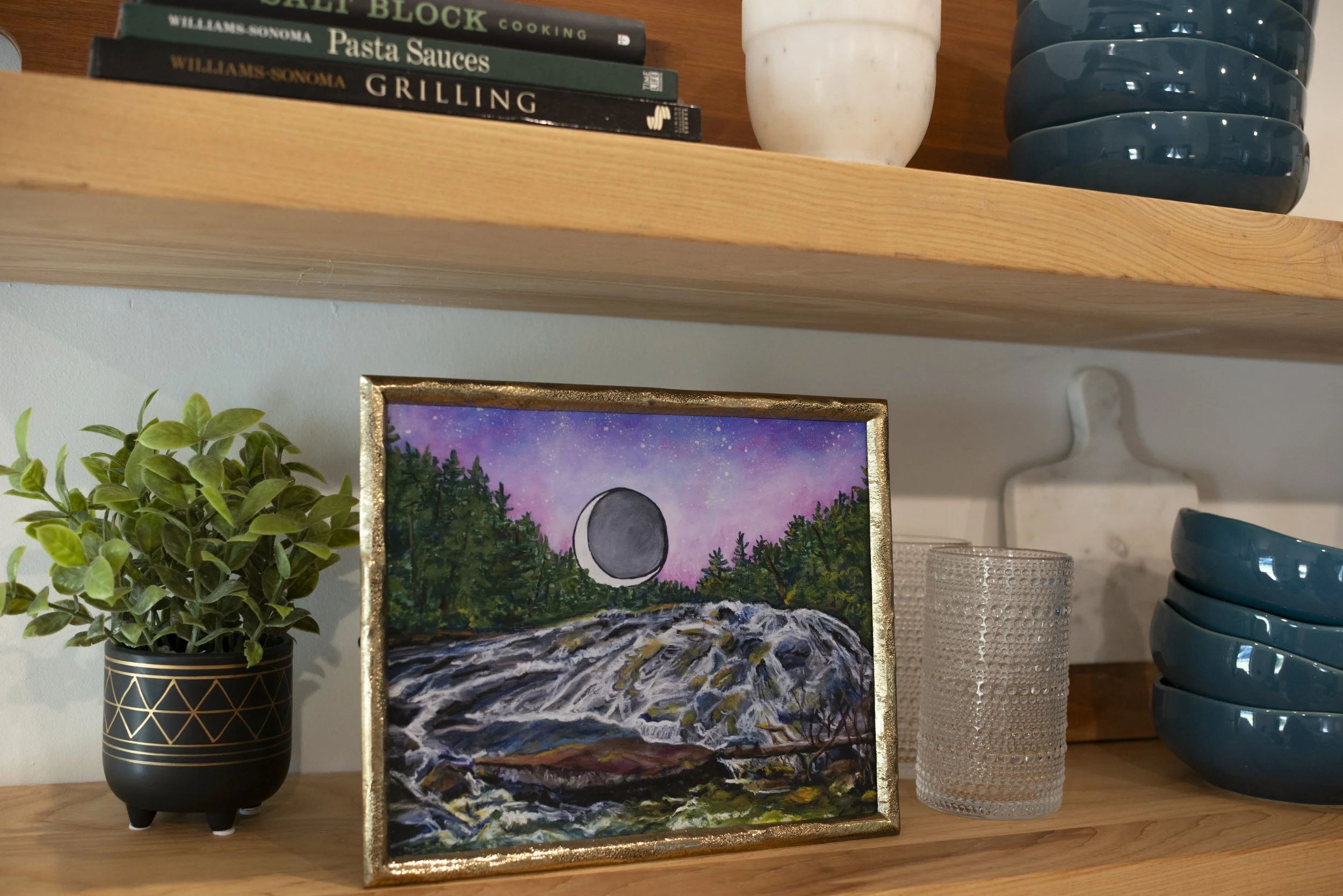 A print of Bridal Veil Falls in DuPont Forest in a gold frame displayed on a shelf with decorative vases, glasses, dish ware, and cookbooks.