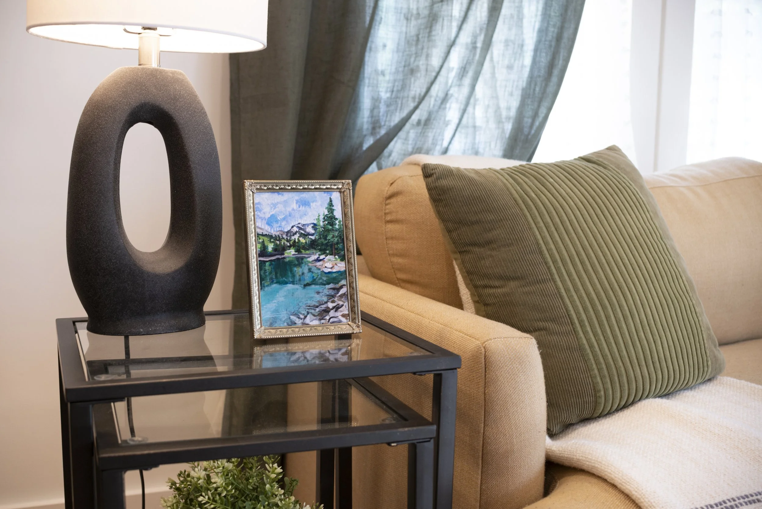 Alpine lake fine art print in a gold frame, placed on a glass living room table with a lamp and houseplant and positioned next to a comfortable couch.
