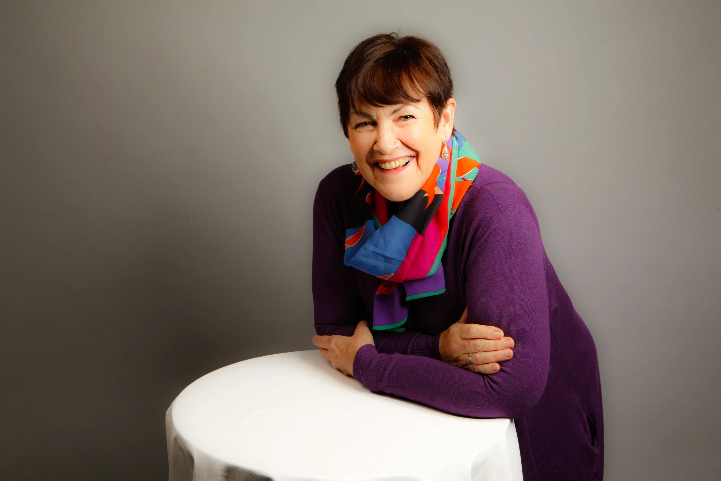 A smiling woman with short brown hair, wearing a purple sweater and a colorful scarf, sitting at a white round table against a plain gray background.