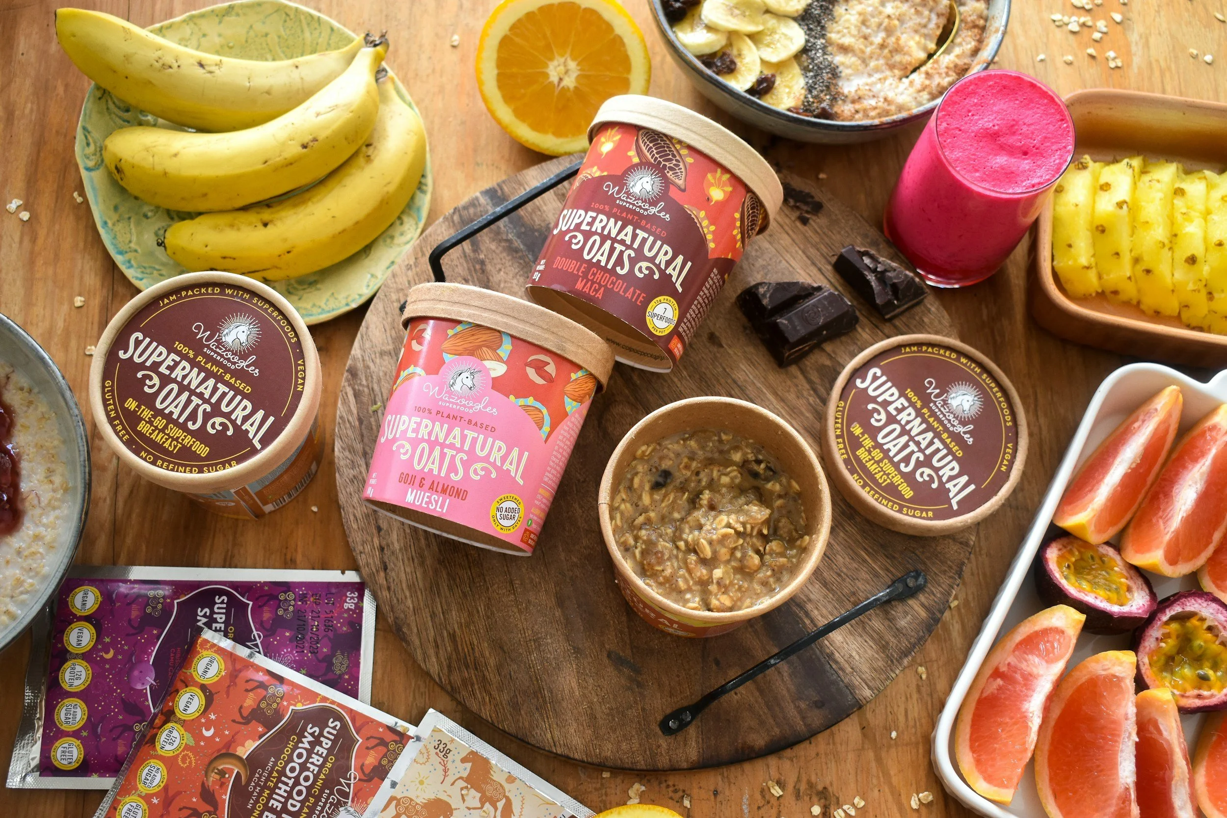 A variety of plant-based foods including bananas, orange slices, oat-based spreads, a pink smoothie, pineapple chunks, and fruit slices on a wooden table.