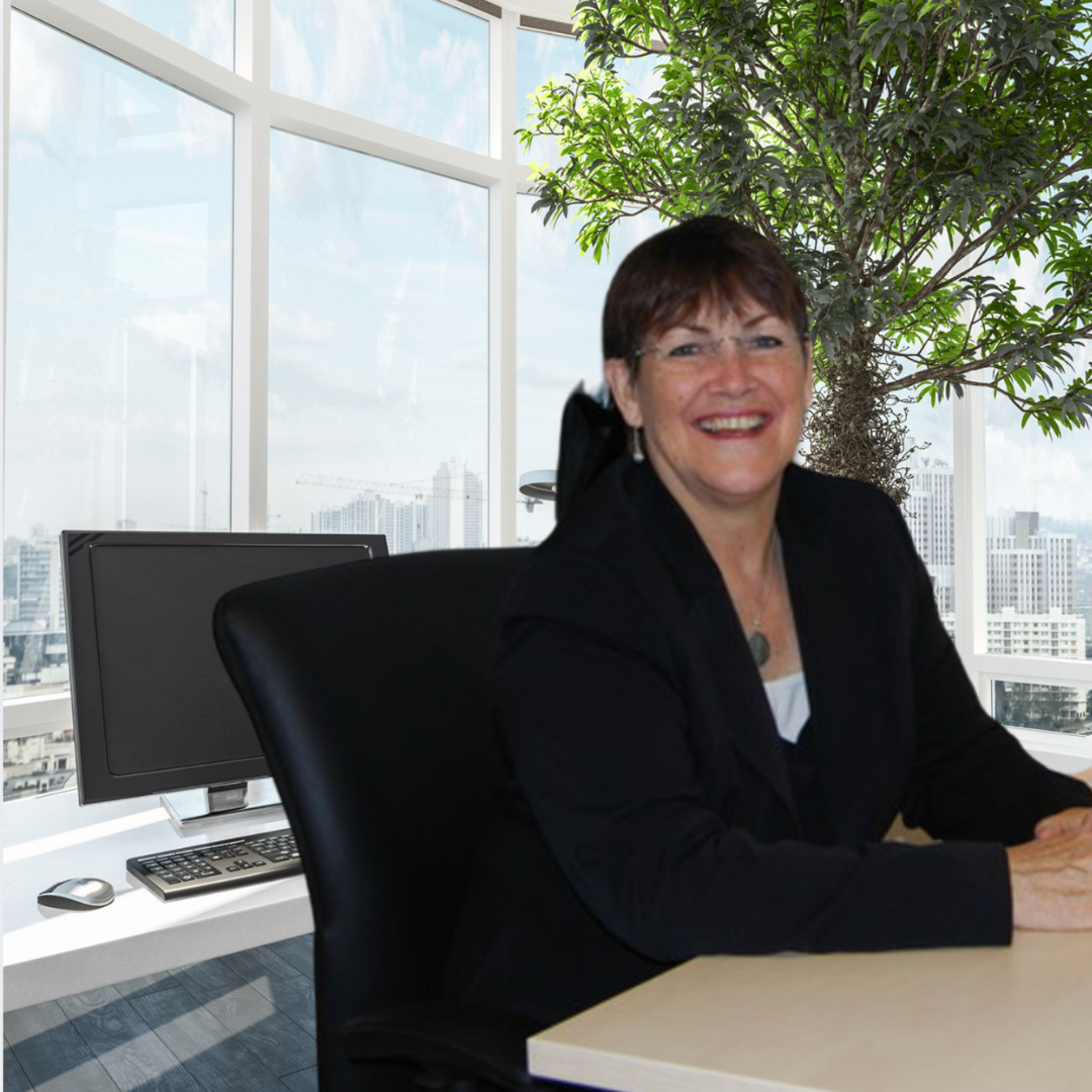 A woman with short brown hair and glasses smiling at the camera, sitting at a desk in a modern office with large windows, a computer monitor, and a large green plant in the background.