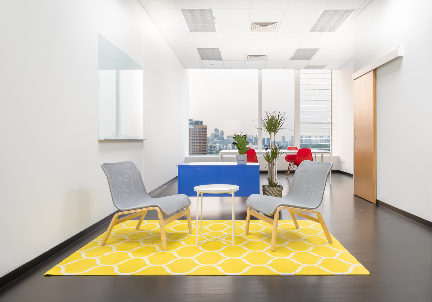 A bright discussion space near the entrance of the office, and formal work stations at the end of the office, divided by a blue cabinet. Immediately to the right of the discussion space is the pantry. The door on the right leads into a private bathr…