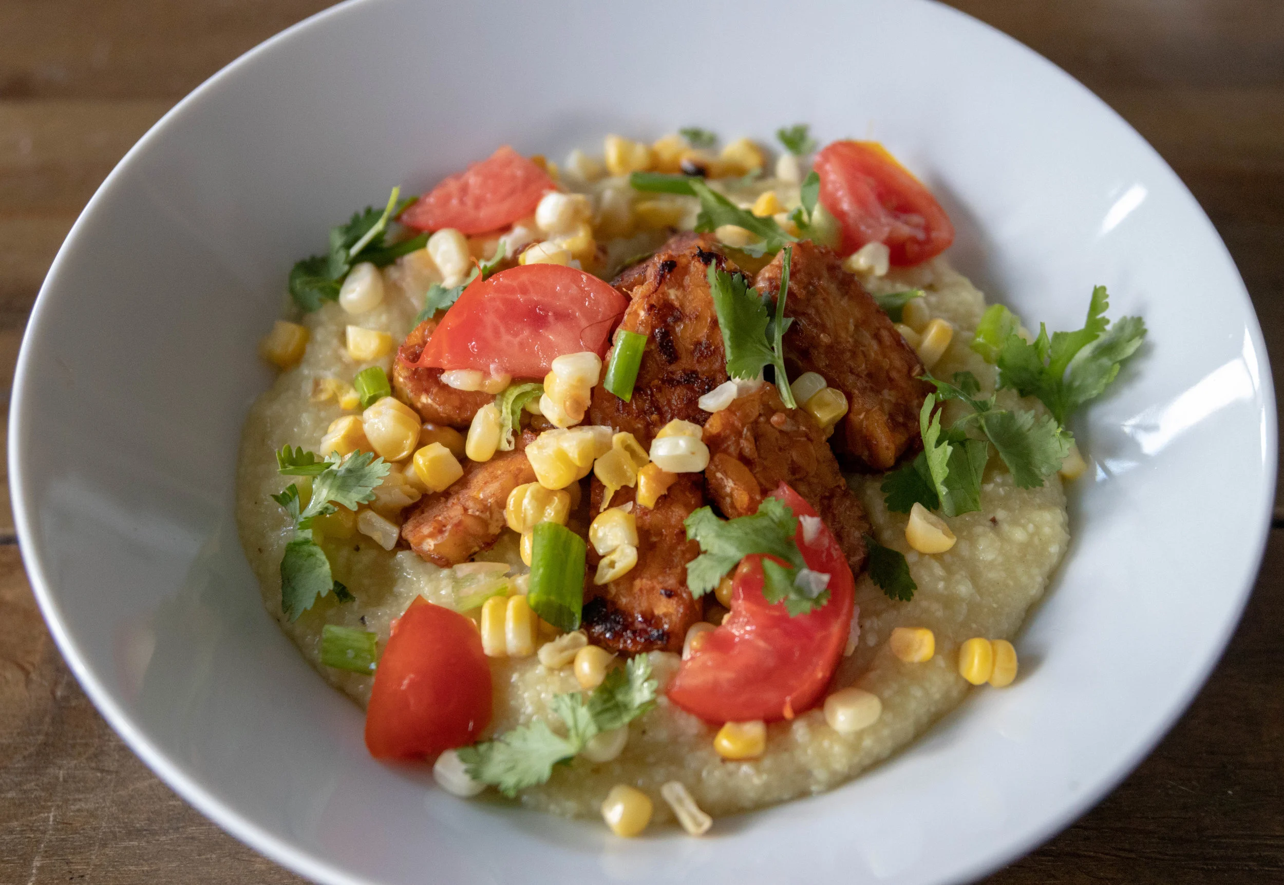 A bowl of grits topped with tempeh, corn, tomatoes, and scallions.