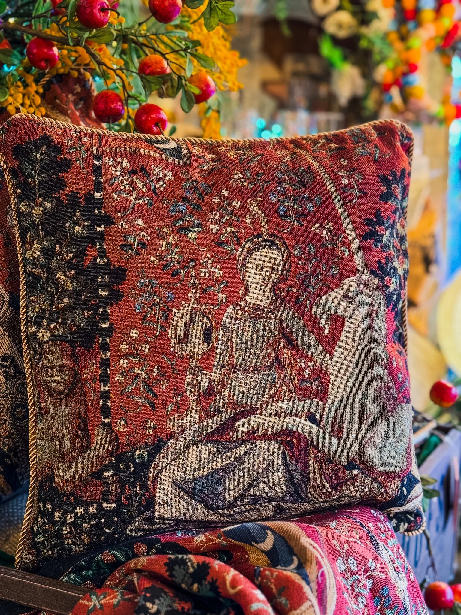 Decorative pillow featuring a tapestry-style image of a woman with a sword, sitting with her eyes closed, amidst floral and nature motifs. The pillow is surrounded by colorful artificial flowers and fruit decorations.