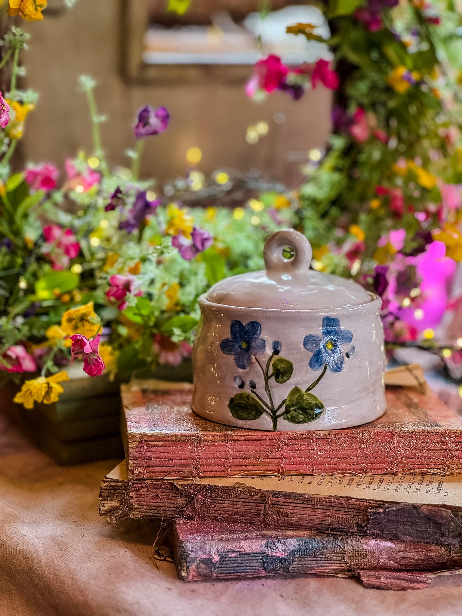 A ceramic container with painted blue flowers sits on a stack of old books with pink covers, surrounded by vibrant pink, purple, and yellow flowers in the background.