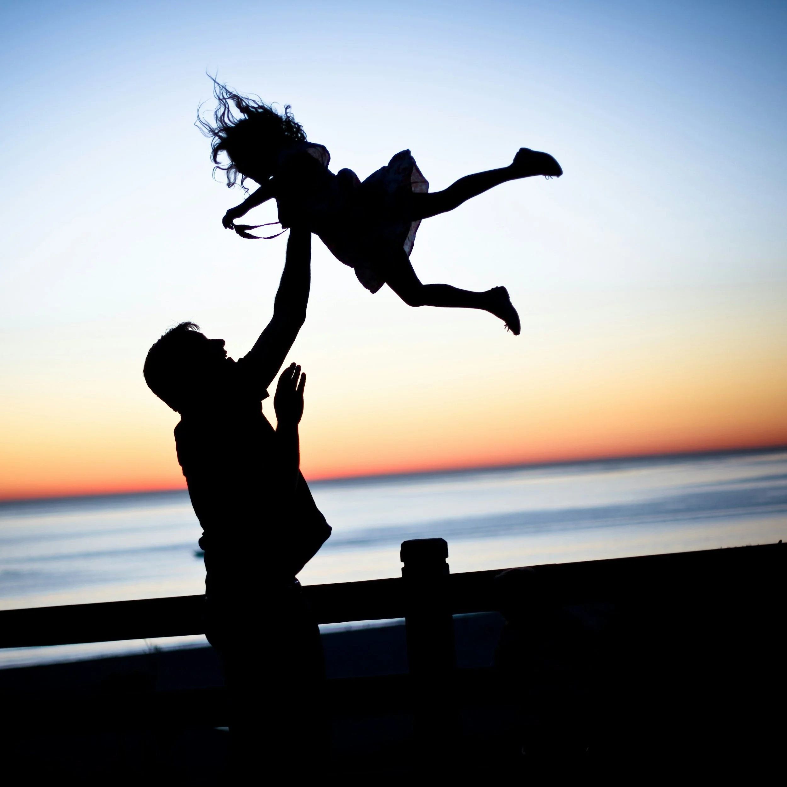 a father plays with her daughter as she yells "more" while he throws her into the air during sunset at the beach, working on her speech therapy targets recently practiced with her speech language pathologist and her father uses strategies he learned