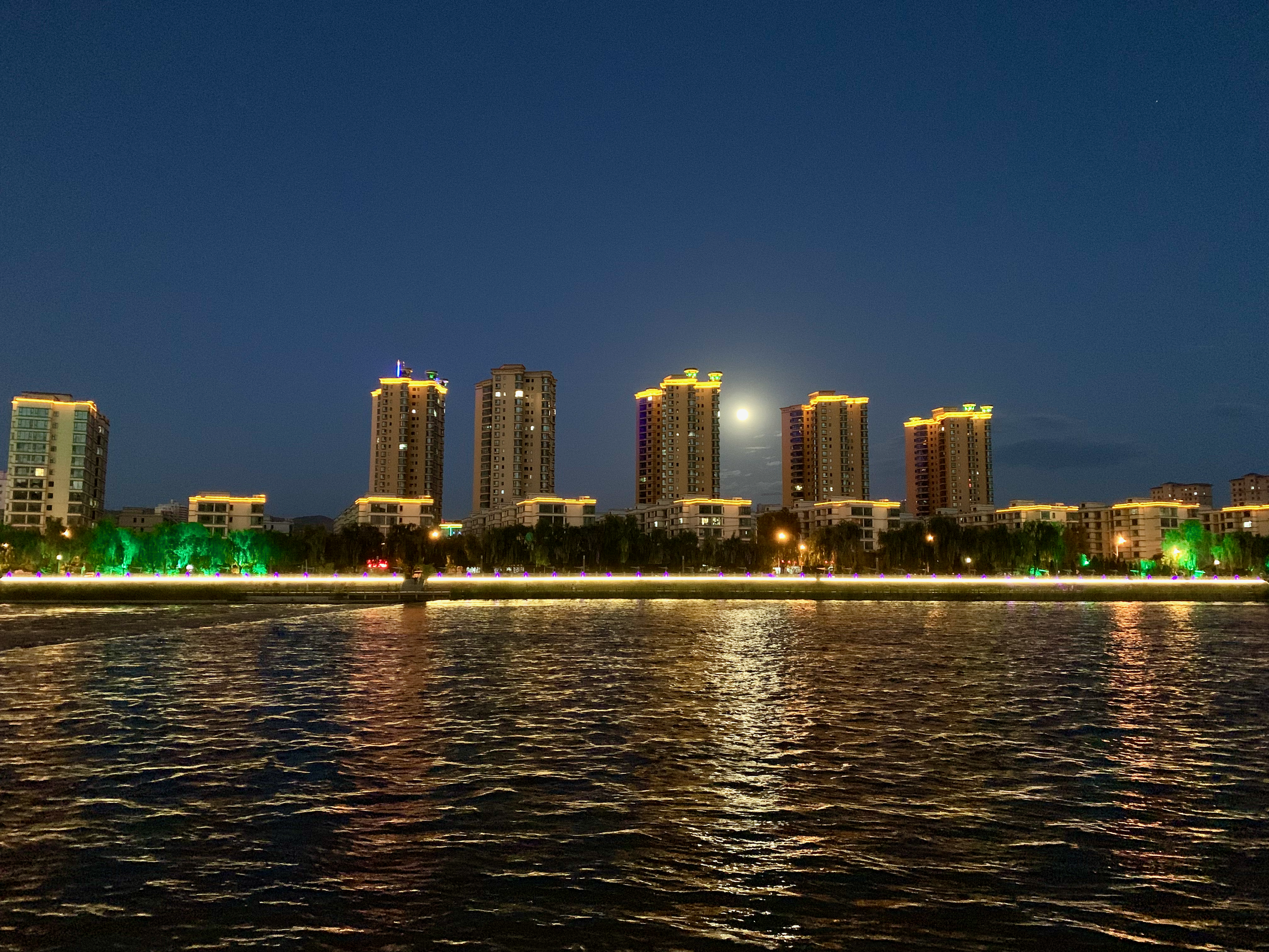 Nighttime cityscape with high-rise buildings illuminated along a waterfront, trees with colorful lights, and a bright moon in the sky.