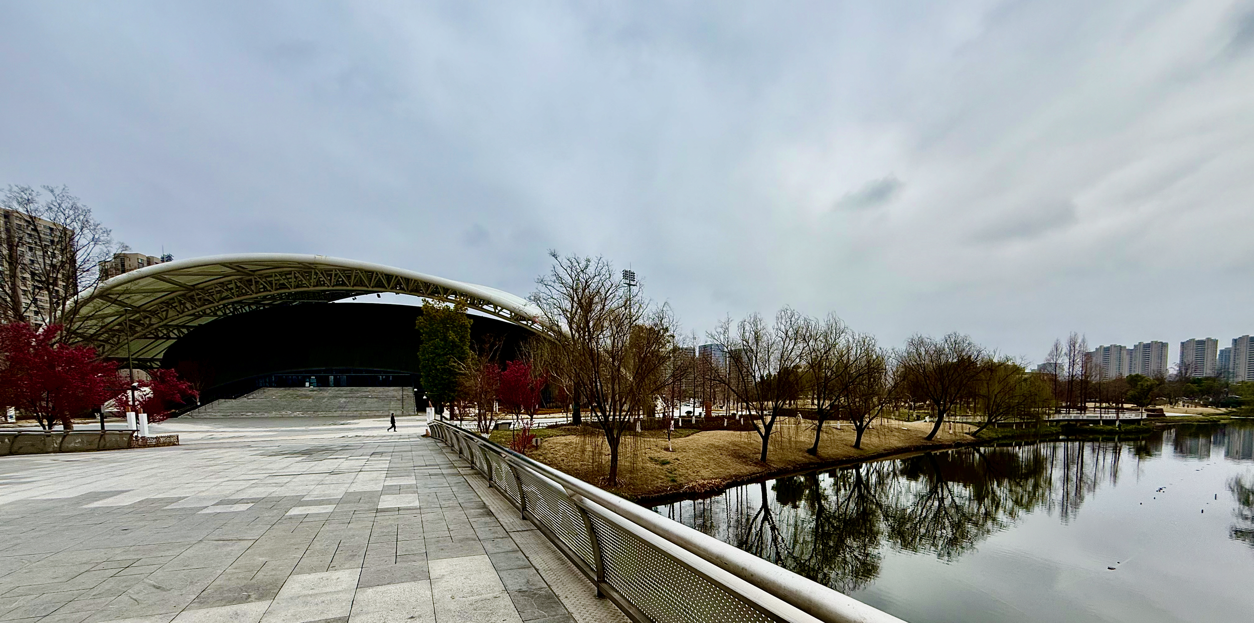 A modern park featuring a large, curved covered stage with a wide, paved pathway and a railing along the water. Leafless trees and reflections in a river are visible, with high-rise buildings in the background under an overcast sky.